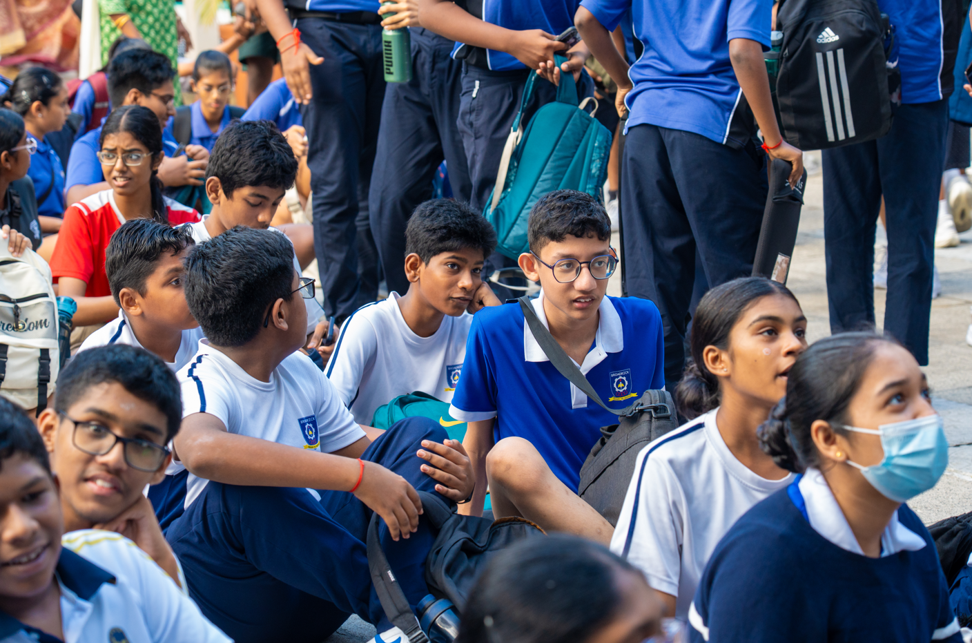 Group of schoolchildren in blue-and-white uniforms sitting on pavement with backpacks, talking.