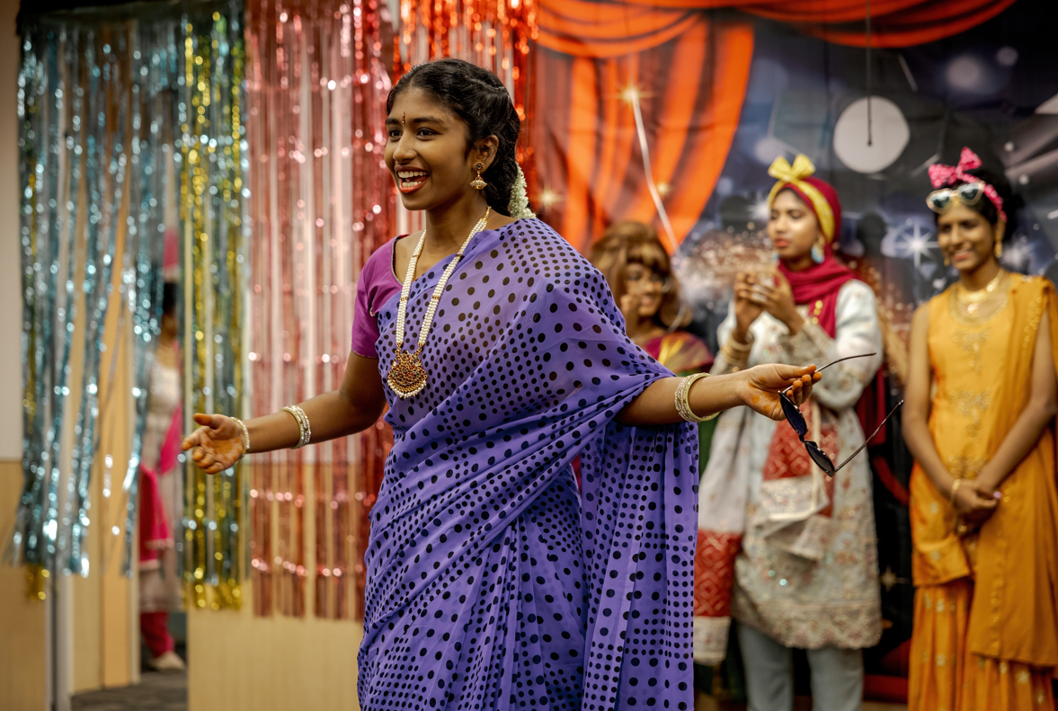 Smiling woman in purple polka-dot sari holds sunglasses, arms out, with people and tinsel backdrop behind.