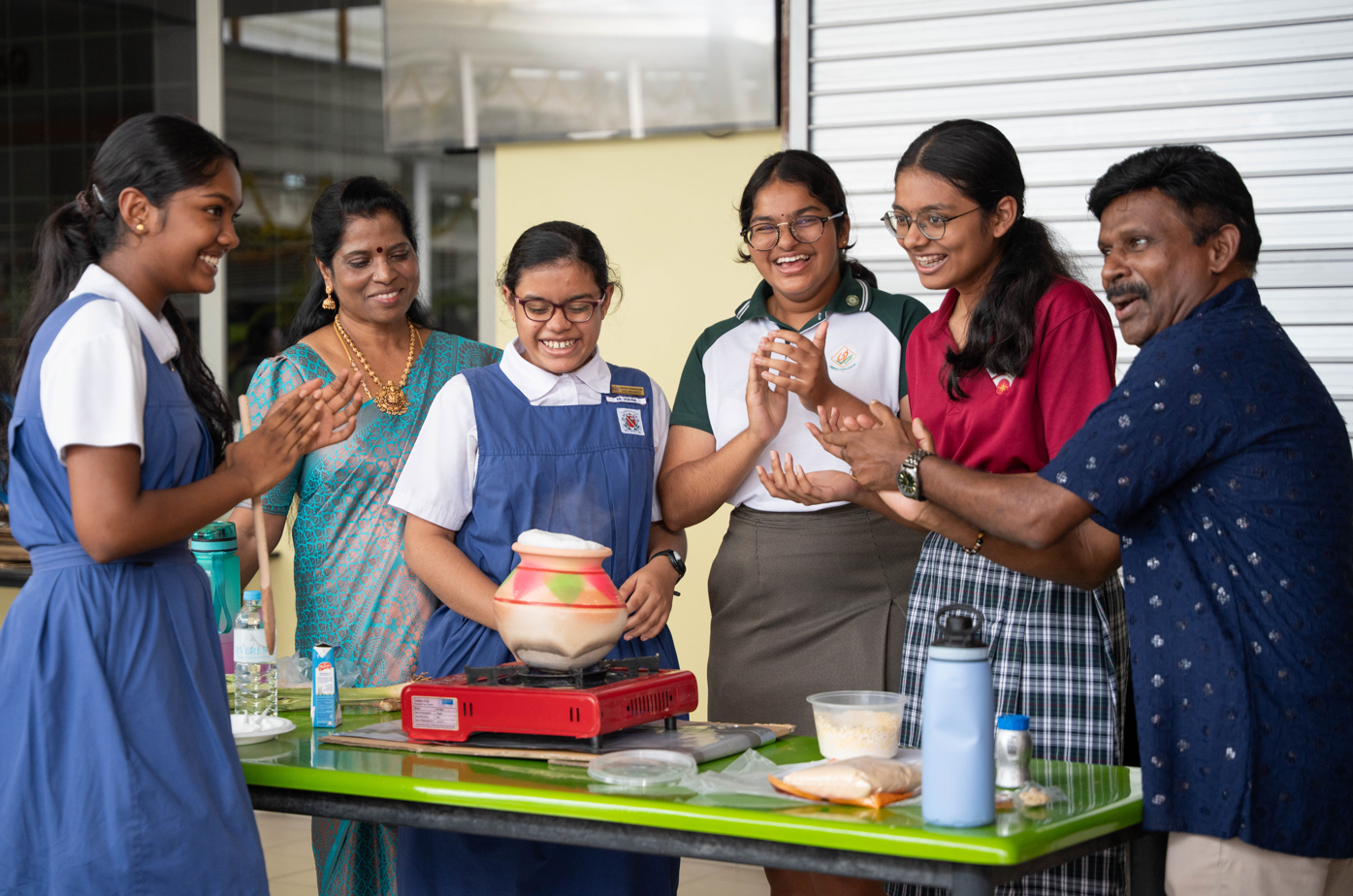 Group of students and adults clap and smile around a table with a pot on a portable stove.