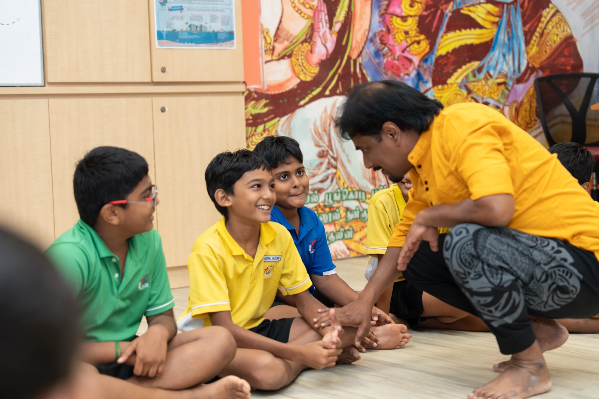 Man in yellow shirt crouches talking to seated boys in colorful uniforms in a room with a mural.