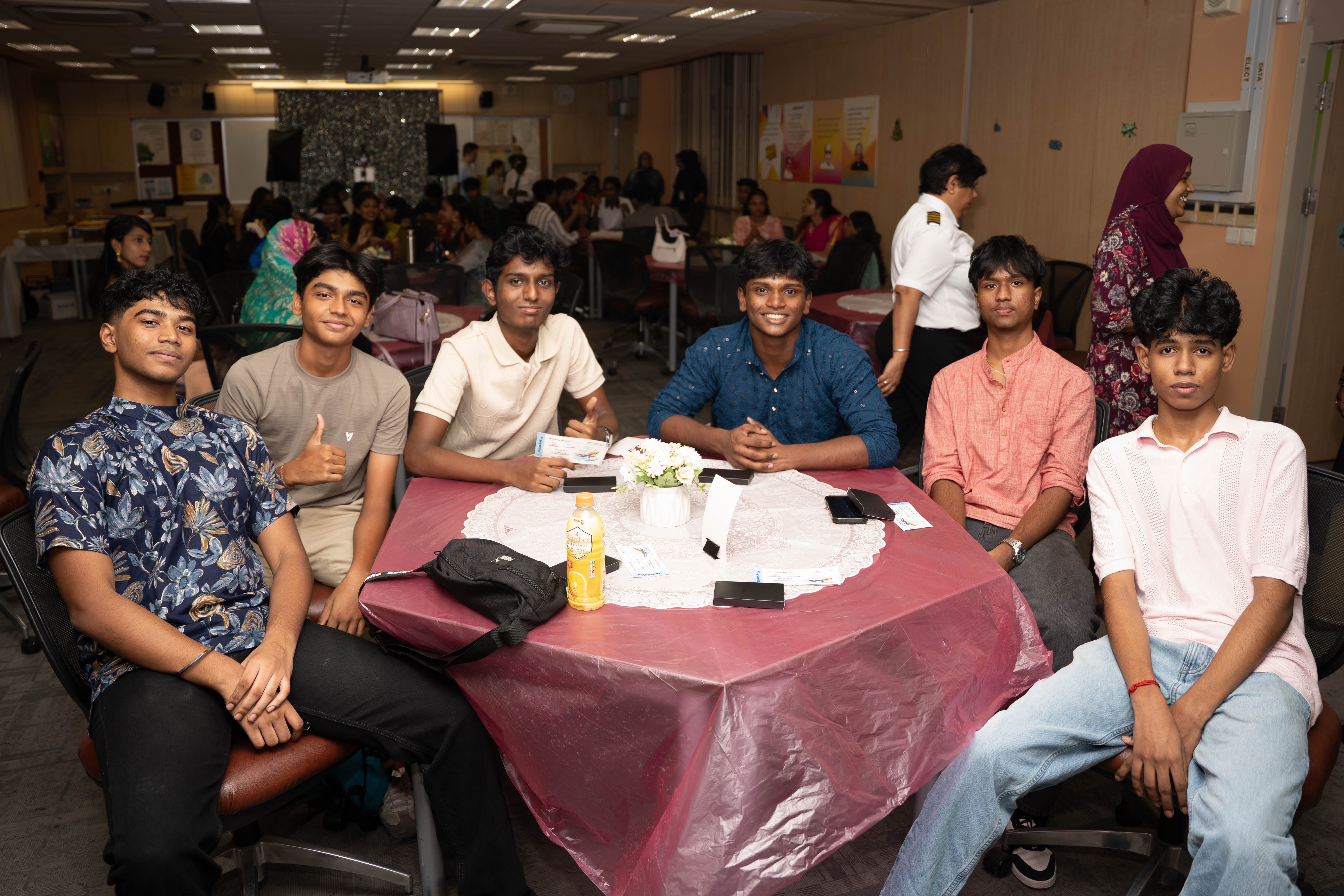 Six young men seated around a table at a busy indoor gathering, smiling toward the camera.