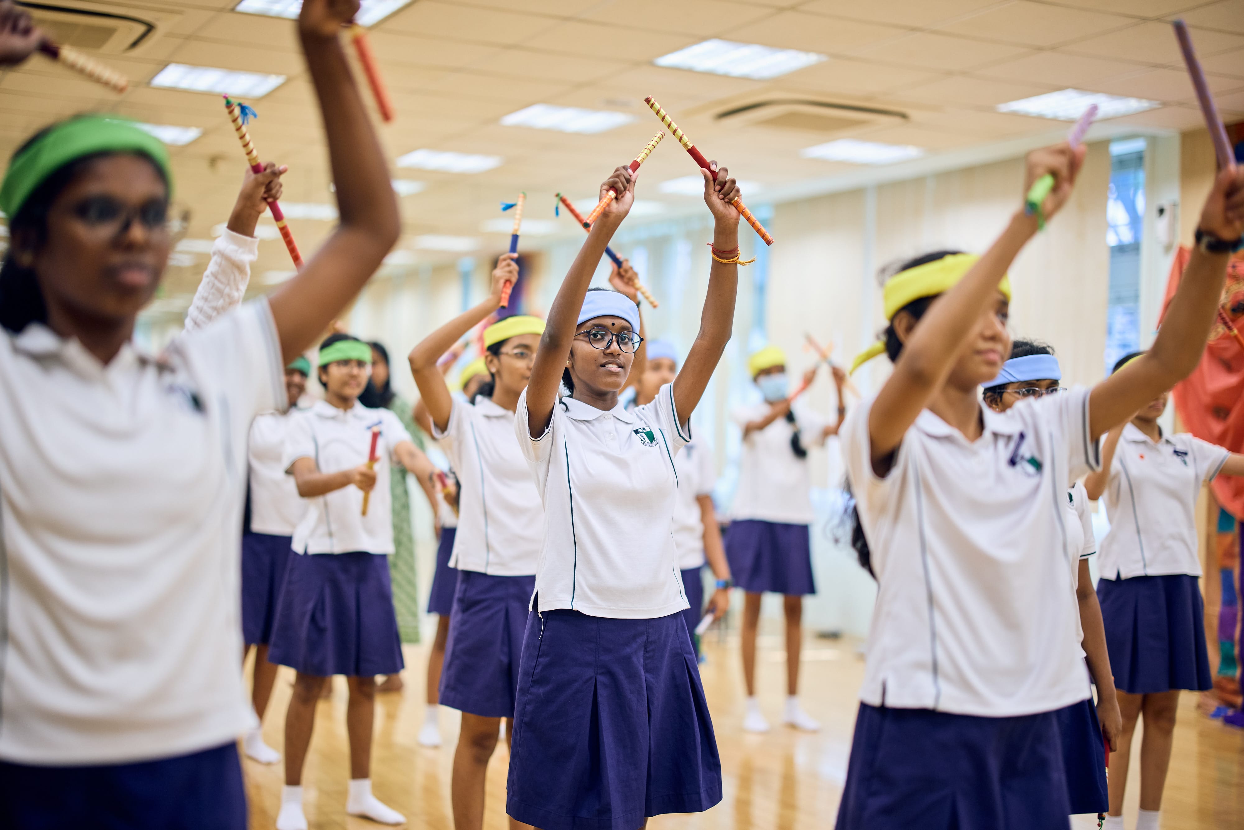 Schoolgirls in white uniforms and blue skirts raise colorful sticks overhead in a bright hall.