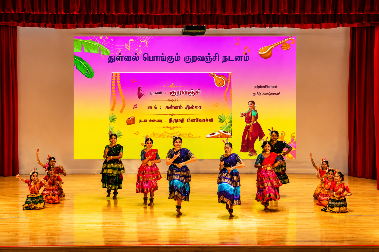 Group of girls in colorful traditional costumes dancing on stage before a large Tamil text backdrop.