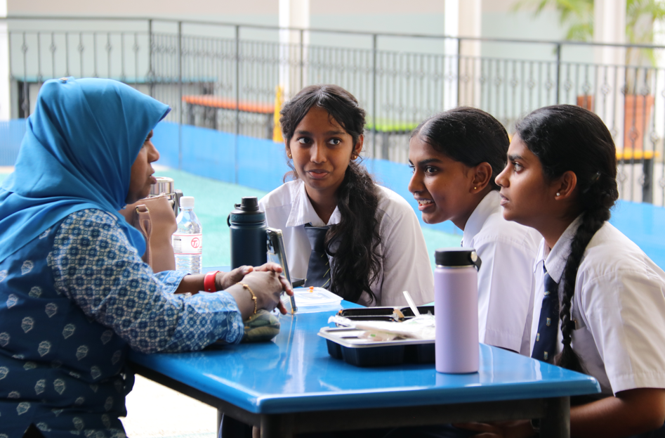 Woman in blue headscarf talks with three schoolgirls in uniforms at a blue table with bottles and tray.