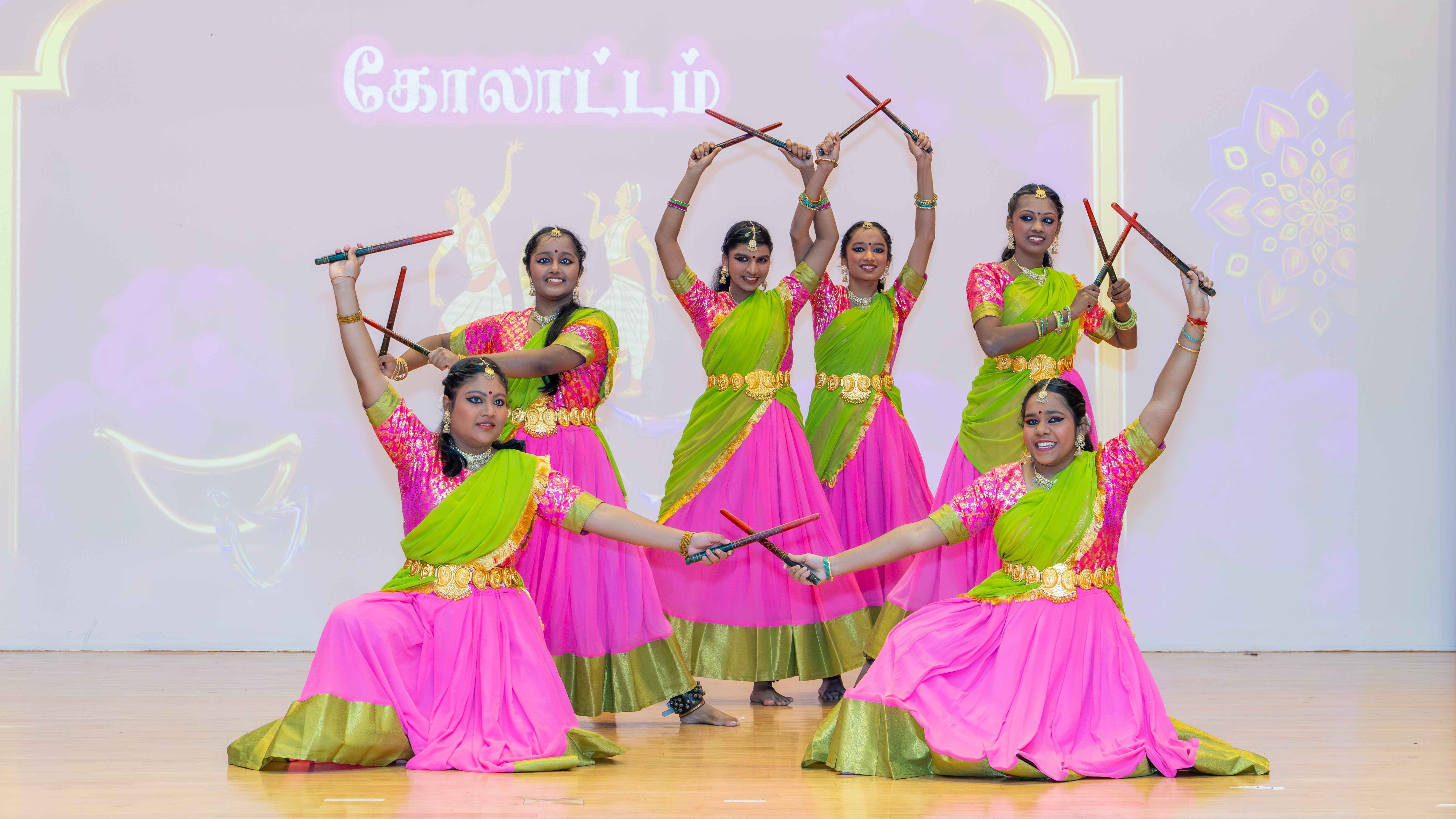 Six dancers in pink and green costumes pose with crossed sticks on a stage backdrop with Tamil text.