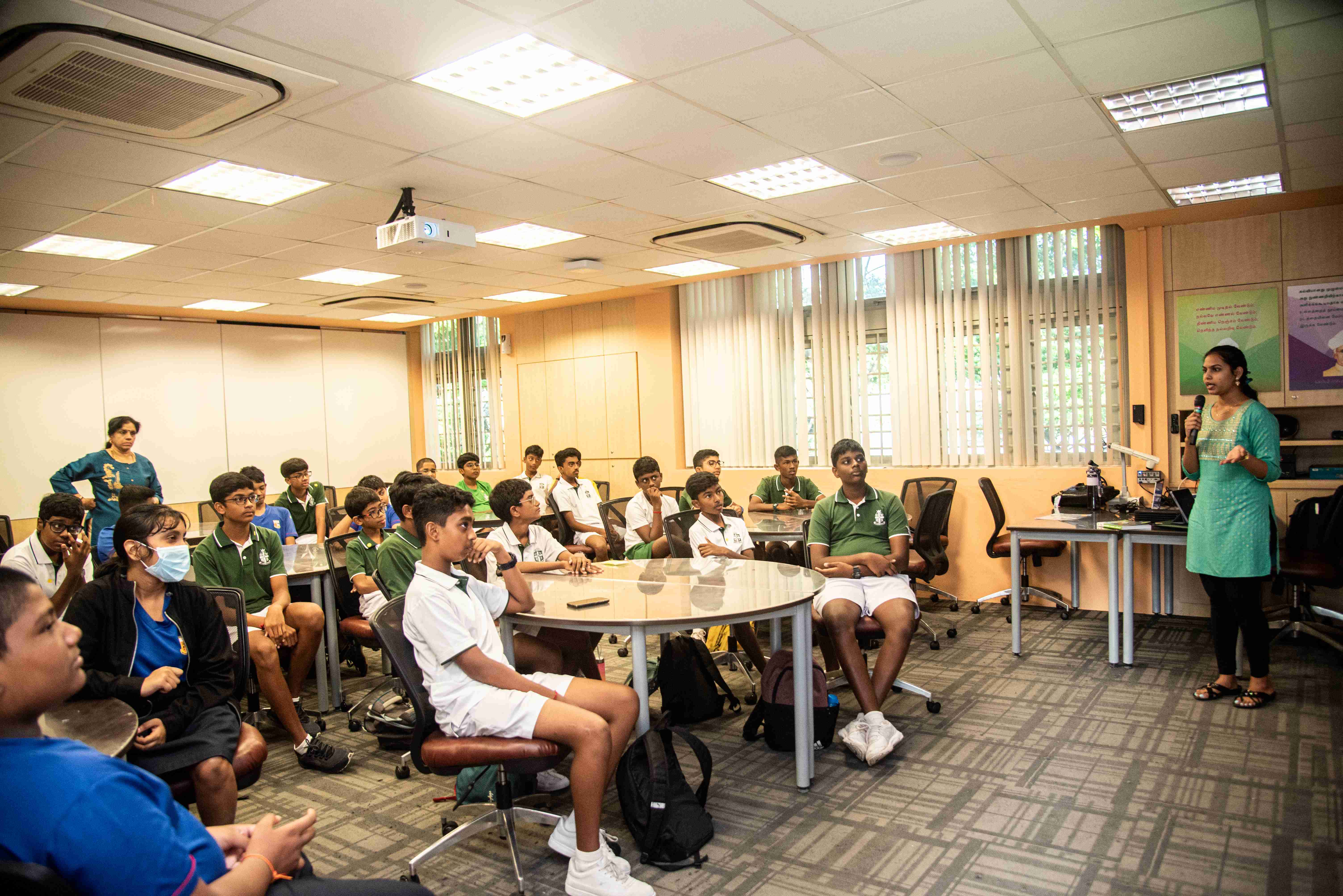 Teacher speaking into a microphone to seated students in uniforms in a classroom with round tables.