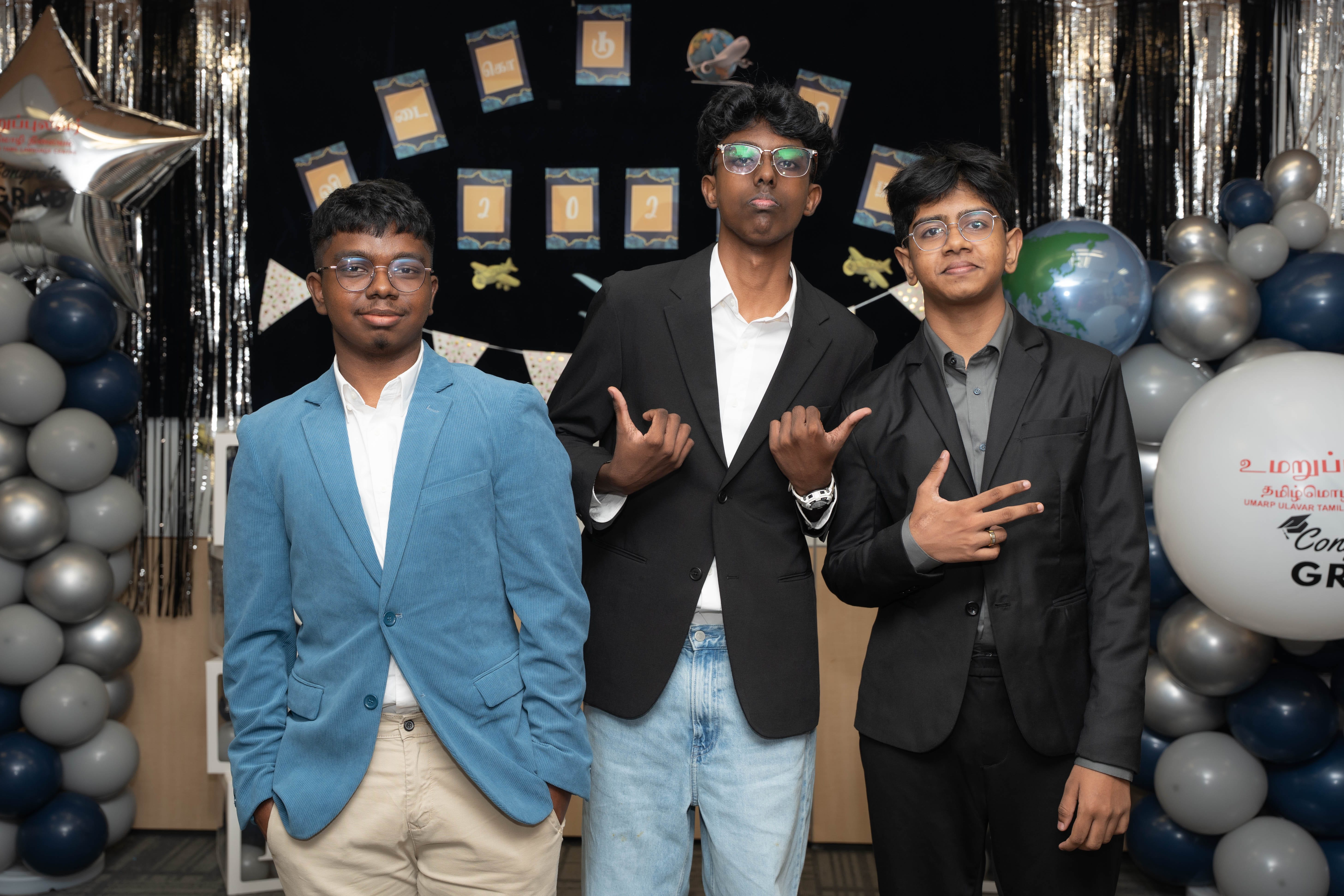 Three young men in suits pose with hand signs before a decorated balloon backdrop and 2025 banner.