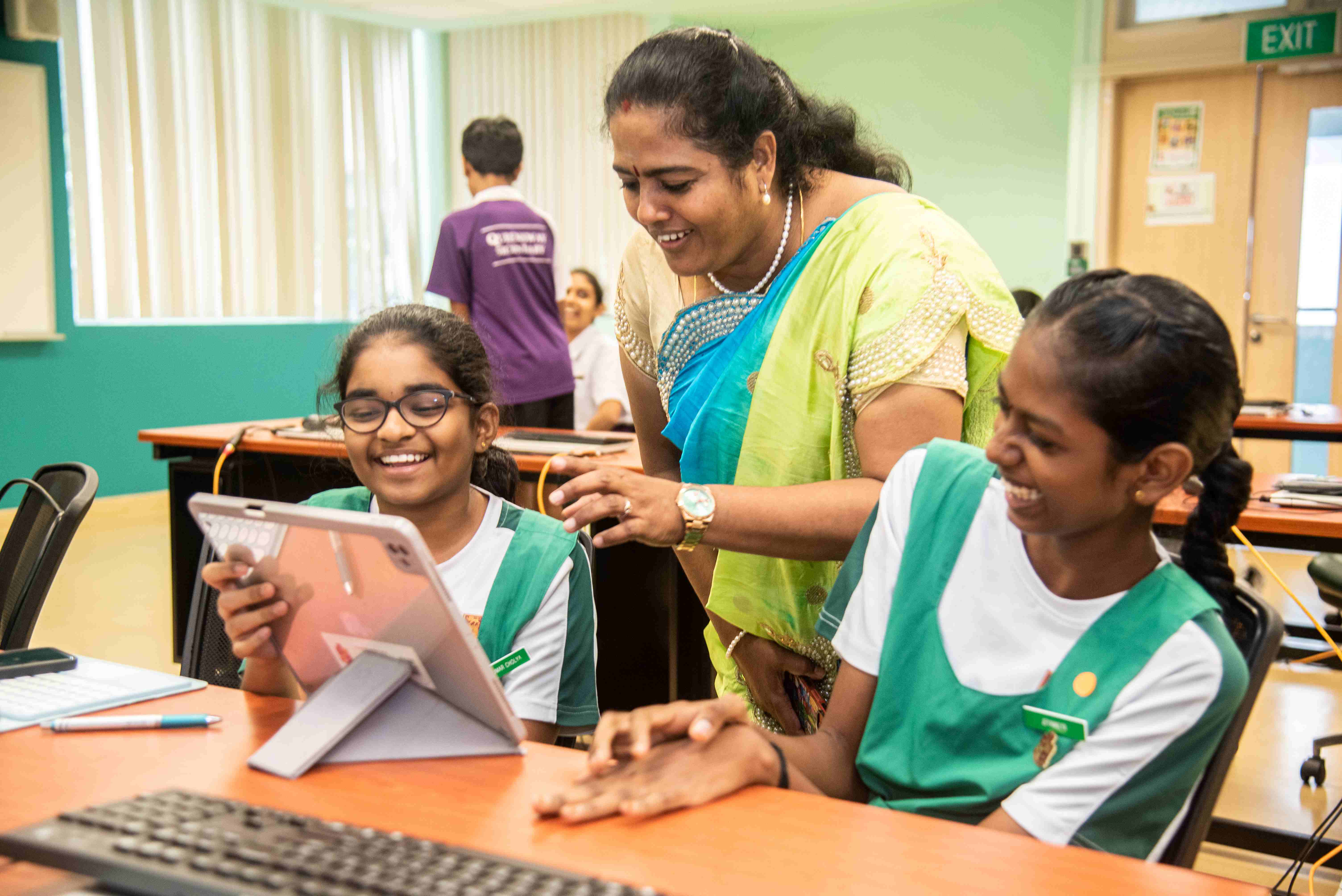 Teacher in a sari smiles with two students in green uniforms using a tablet at a classroom desk.