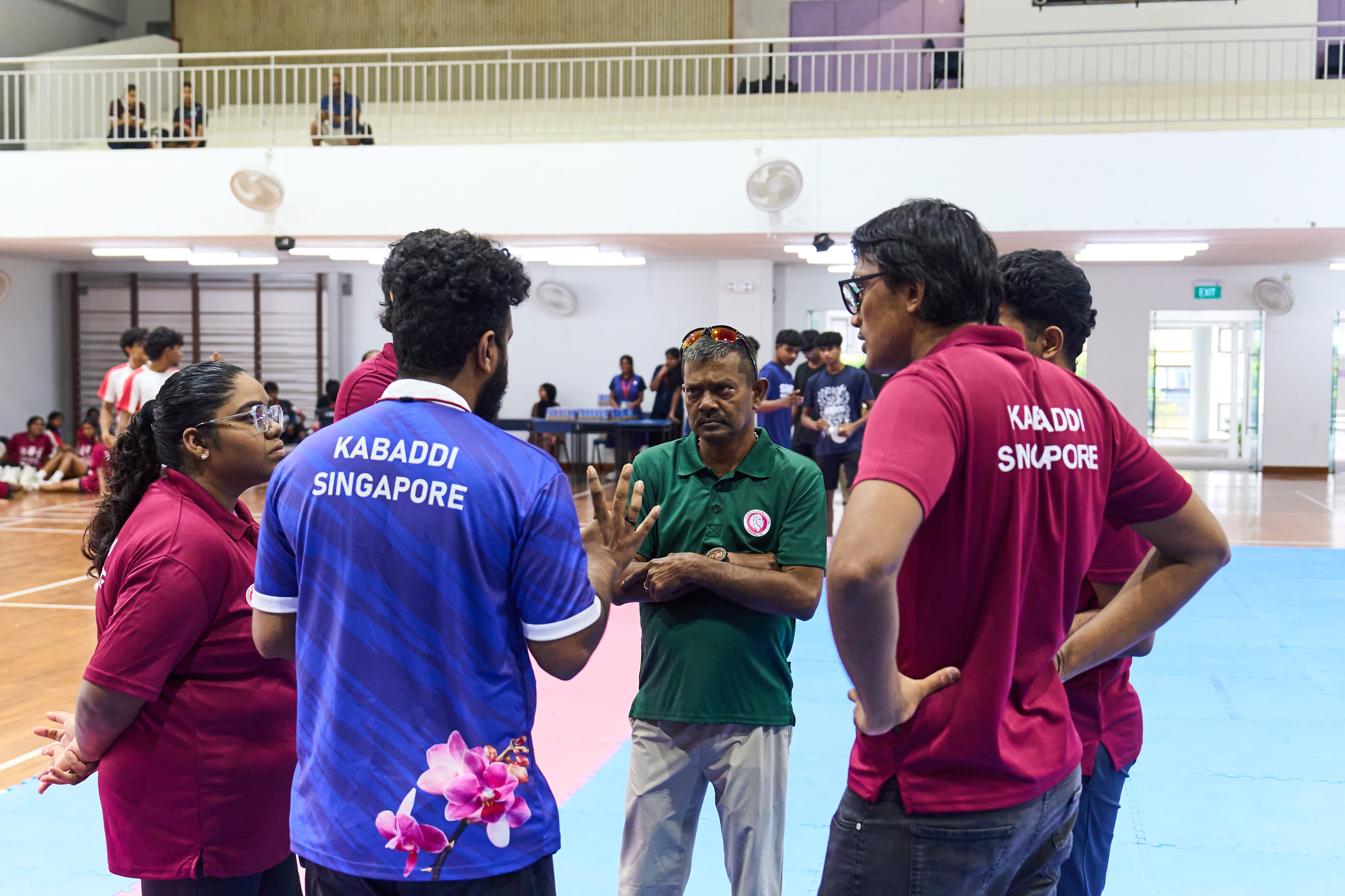 Group of coaches in Kabaddi Singapore shirts talk with a referee on an indoor court.