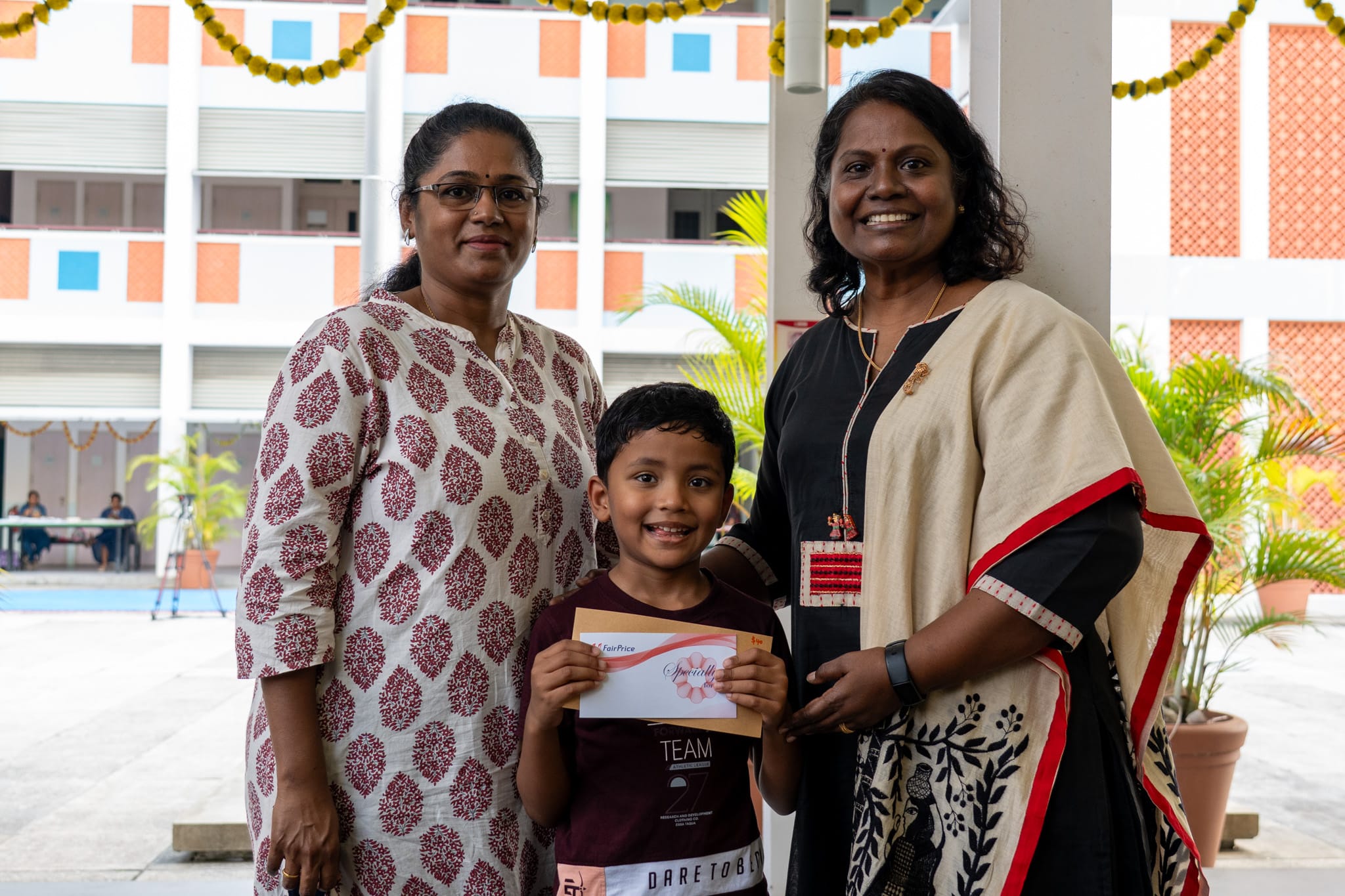 Two women stand with a smiling boy holding a certificate, in a decorated school courtyard.