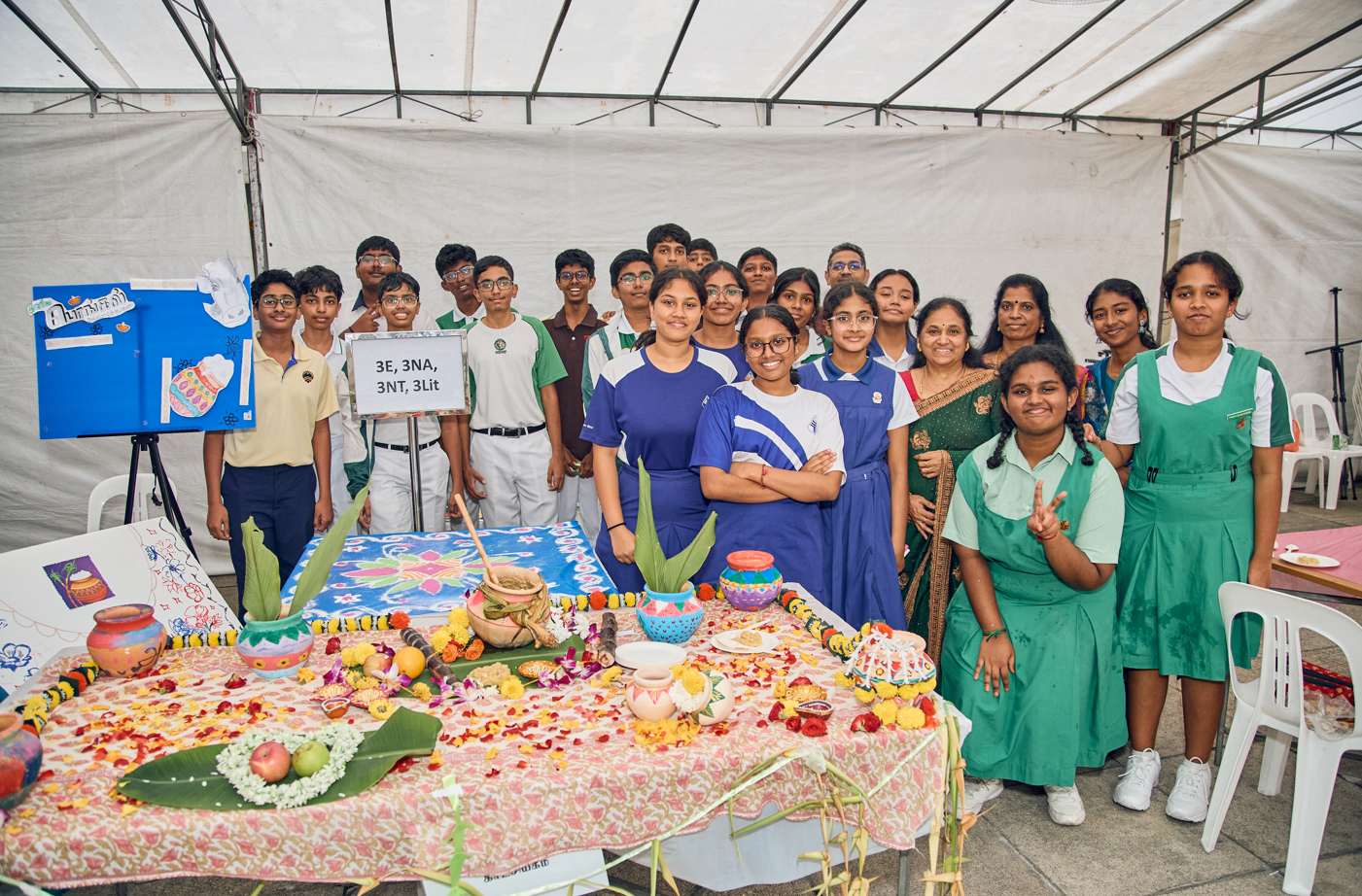 Group of students and teachers pose behind a decorated table with flowers, fruits, and painted pots under a tent.