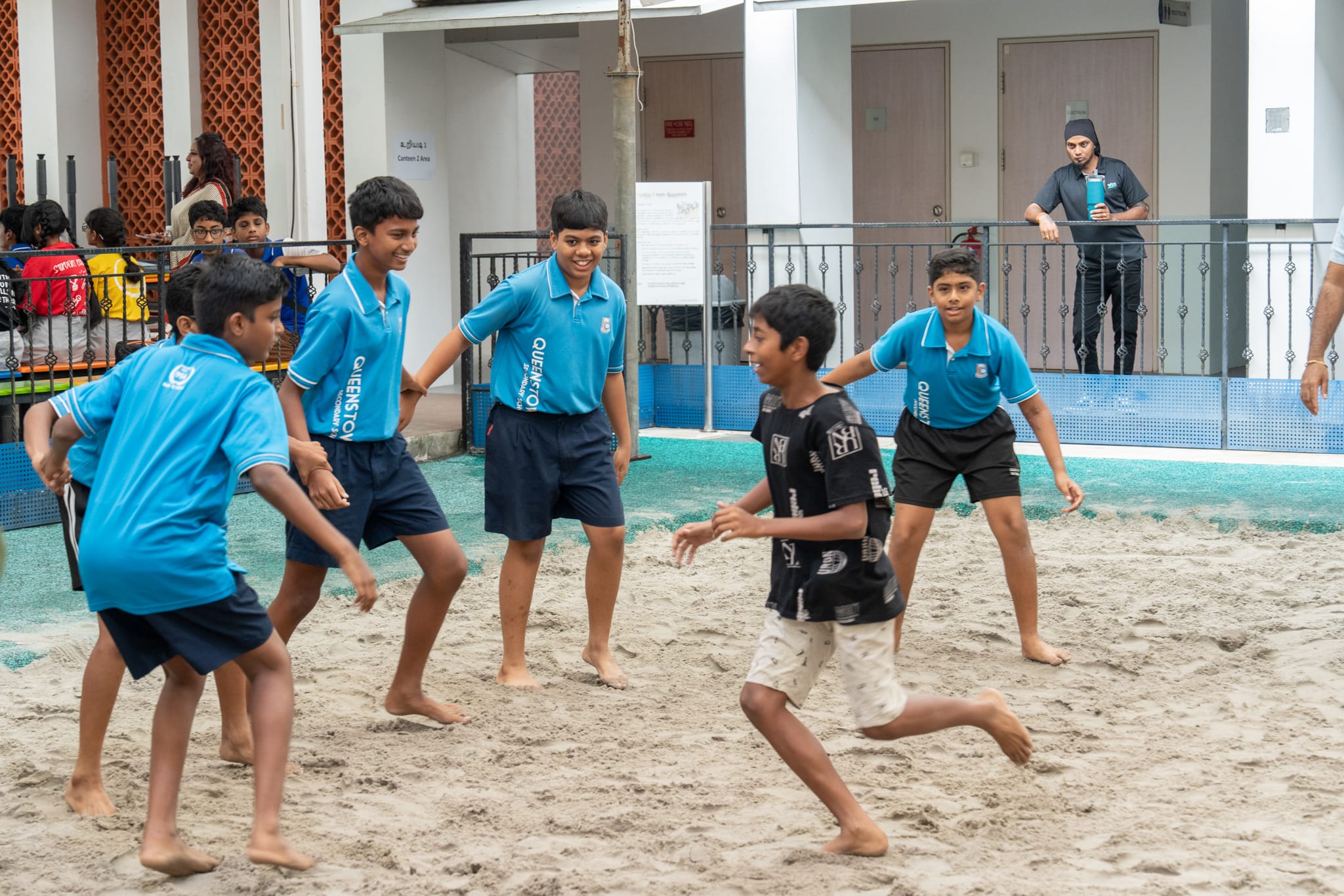Boys in blue uniforms play tag on a sandy court as one boy in a black shirt runs past.
