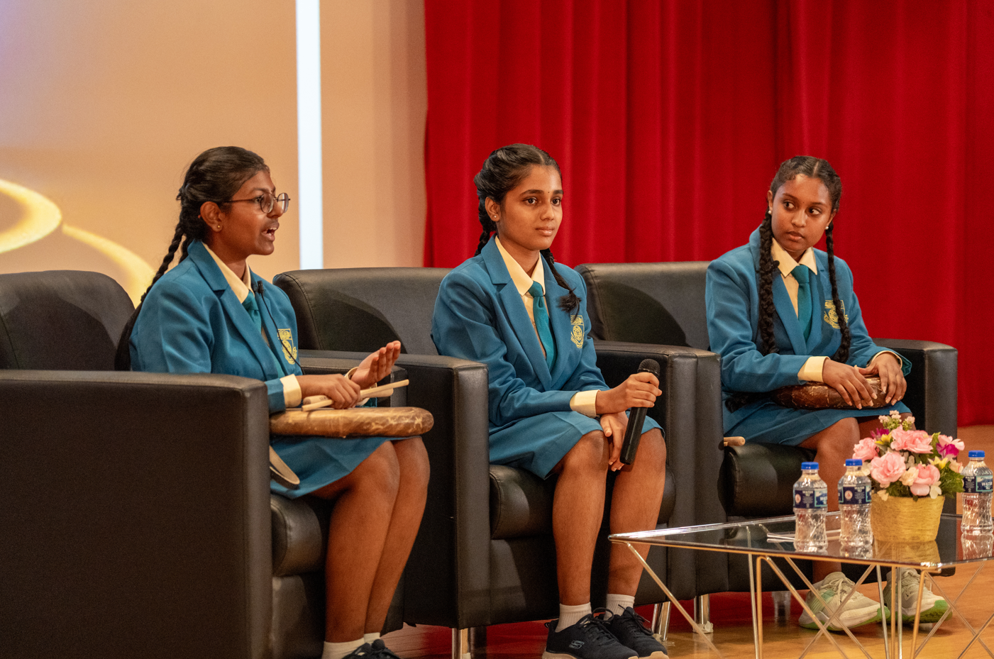 Three schoolgirls in blue uniforms sit in armchairs on stage; one holds a mic, red curtain behind.