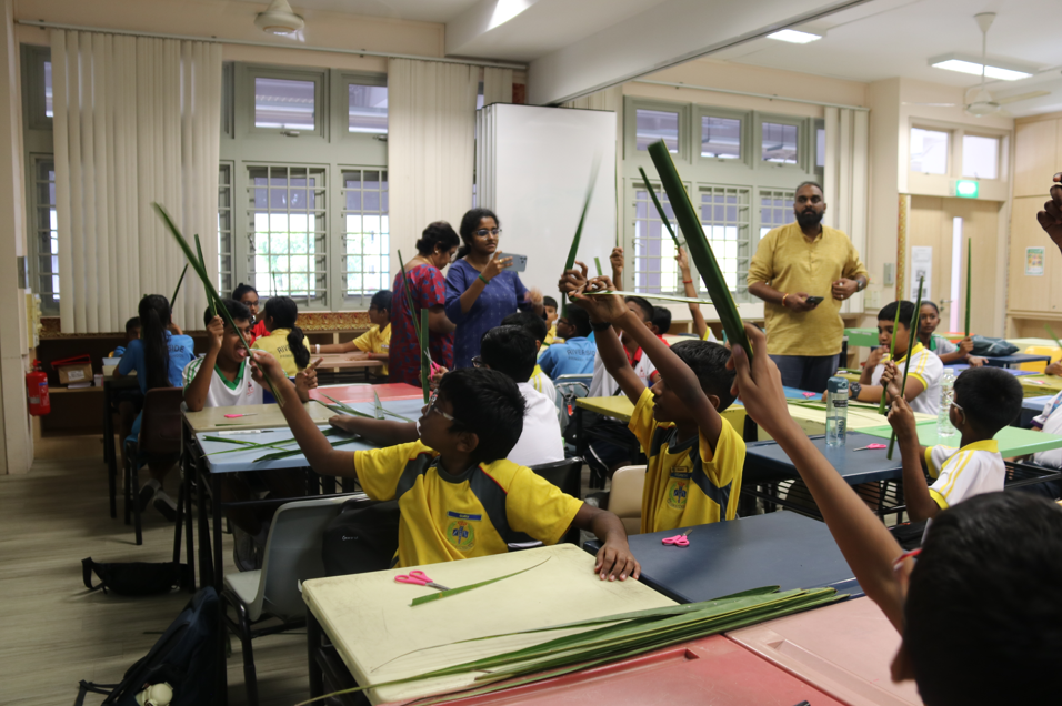 Classroom of children in yellow uniforms raising long green leaves as teachers stand among desks.