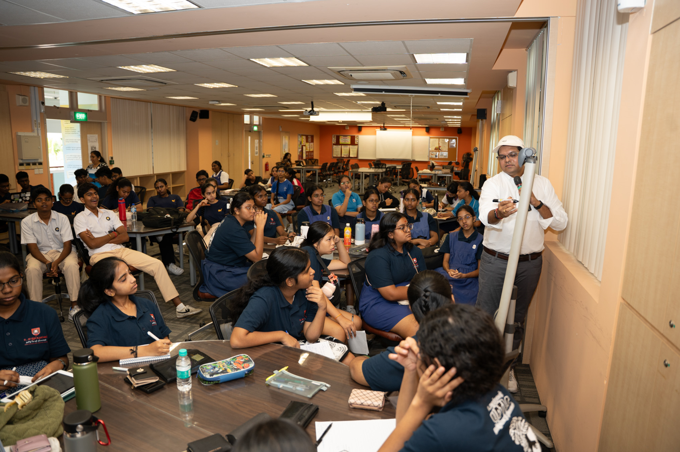 Teacher stands by a screen with a device as students in uniforms sit at tables in a classroom.