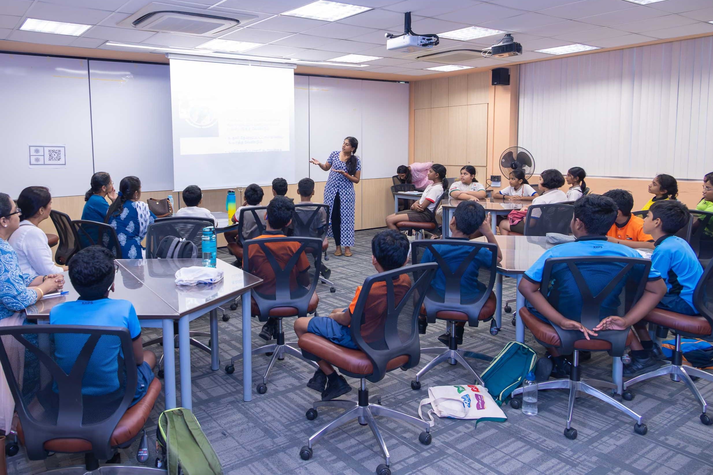 Woman presents at a projector screen while children sit in rolling chairs around tables in a classroom.