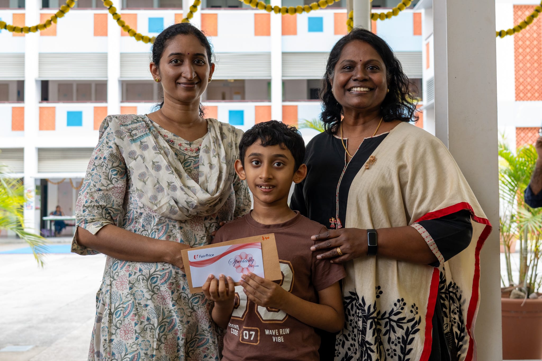 Two women stand beside a boy holding an envelope, all smiling in a decorated school courtyard.