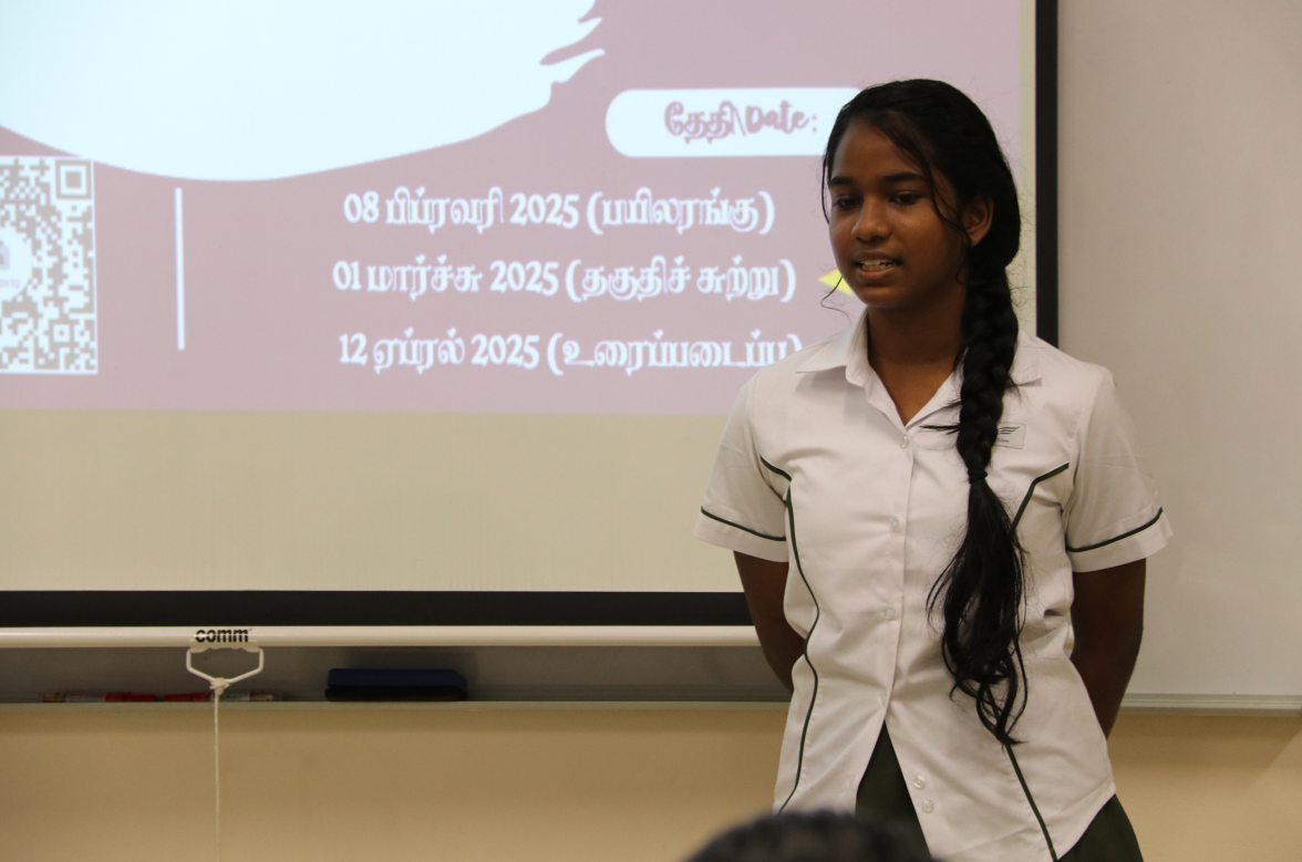Student in white uniform stands before a projected Tamil slide with dates and a QR code.
