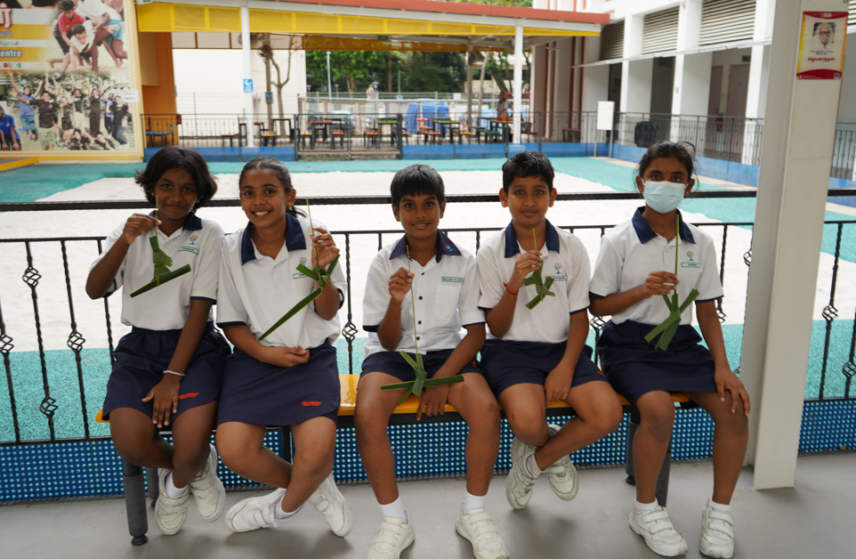 Five school children in white and navy uniforms sit on a bench holding green woven palm leaf crafts.