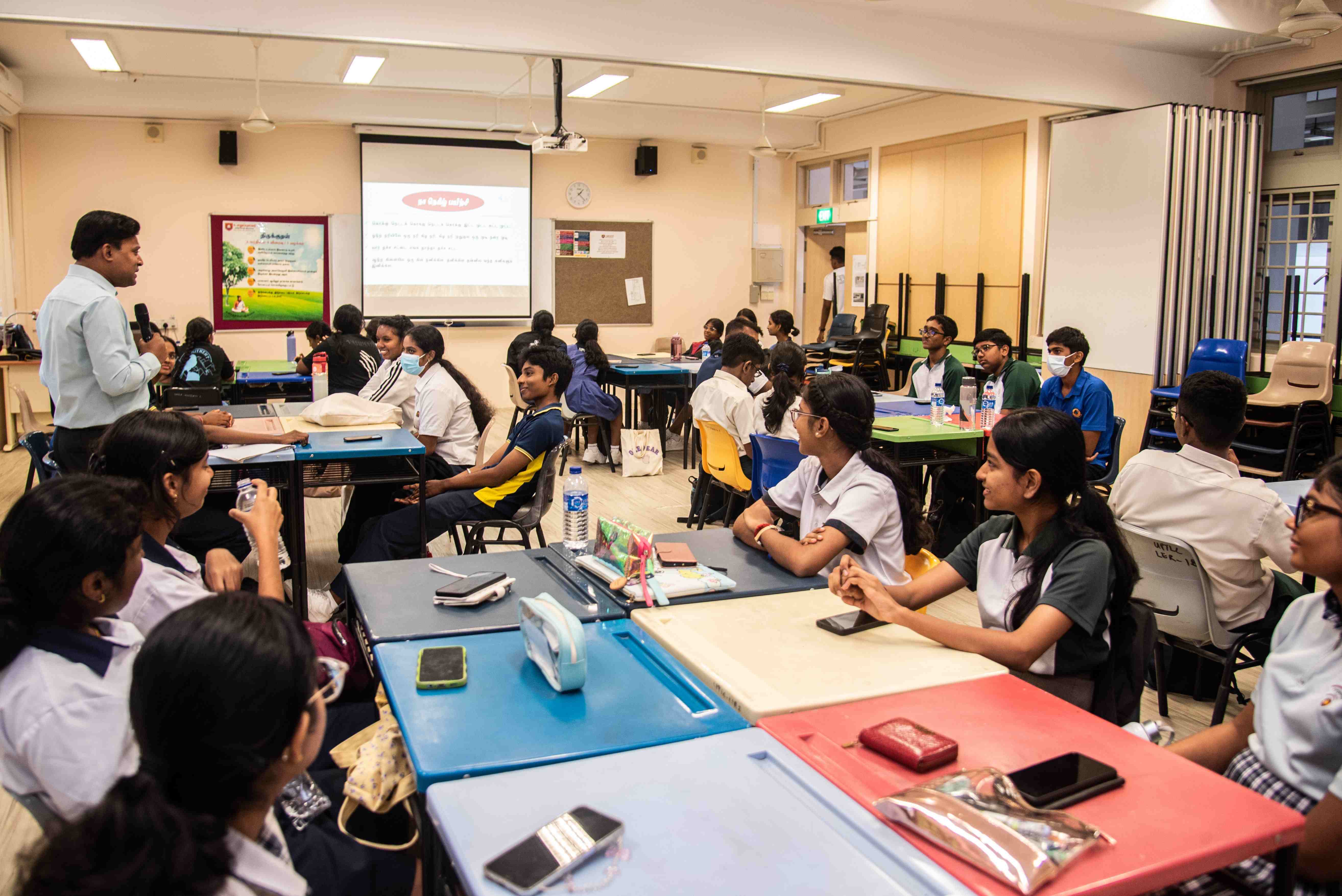 Teacher with microphone addresses students seated at grouped tables in a classroom with a projected slide.