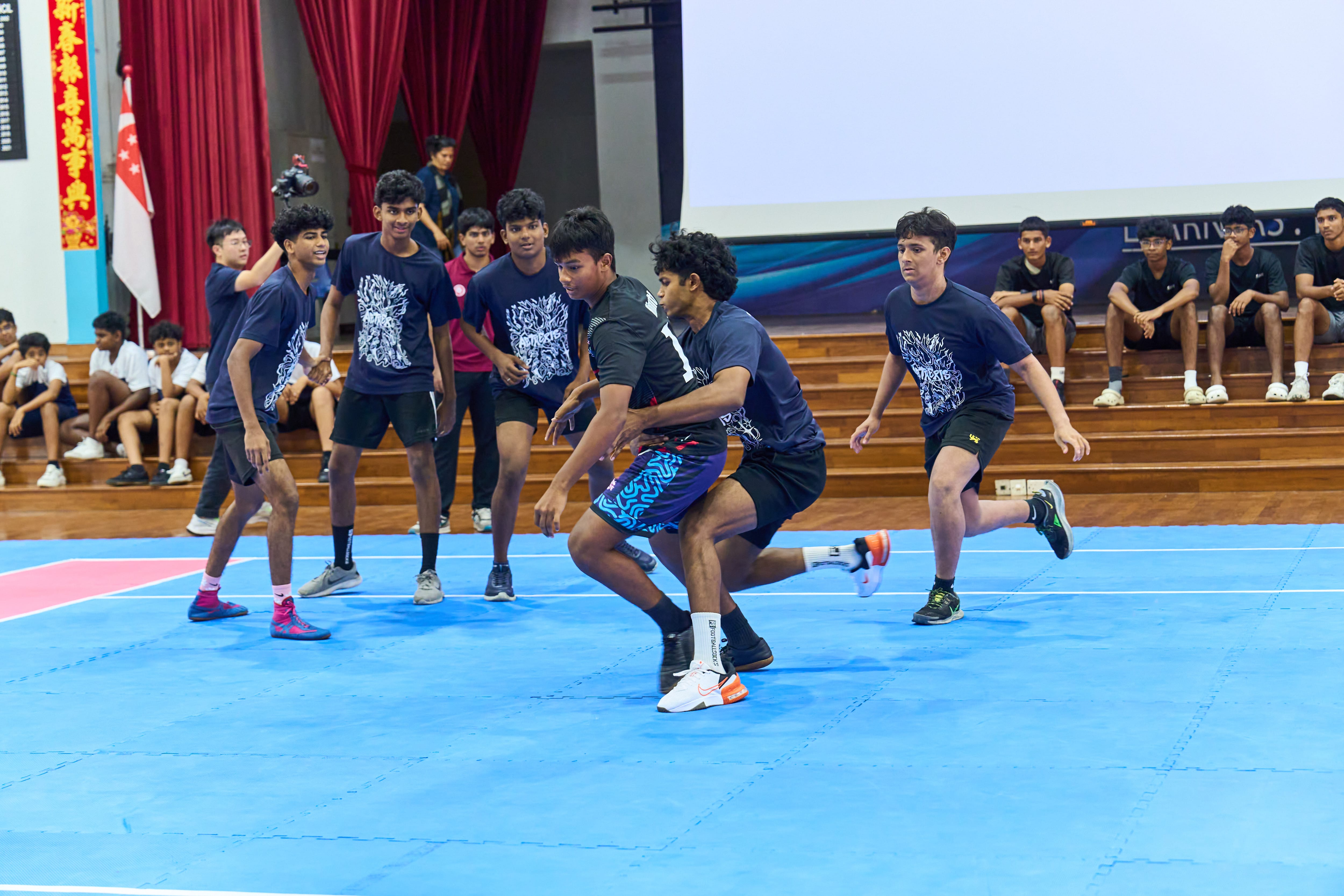 Teens play a sport on blue mats indoors, one grabs another as teammates and spectators watch on bleachers.