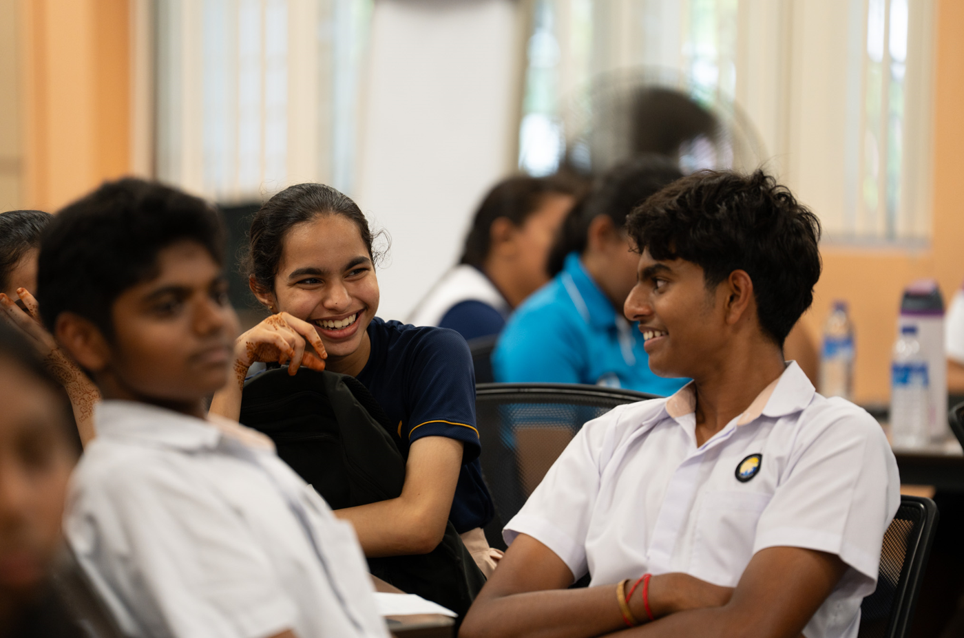 Smiling students chat in a classroom; girl in navy shirt laughs at boy in white uniform.