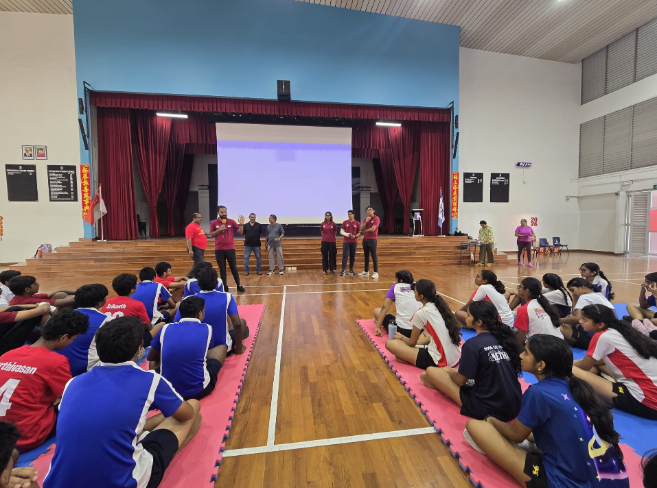 Students sit on mats facing staff speaking on a gym stage with red curtains and a large projection screen.