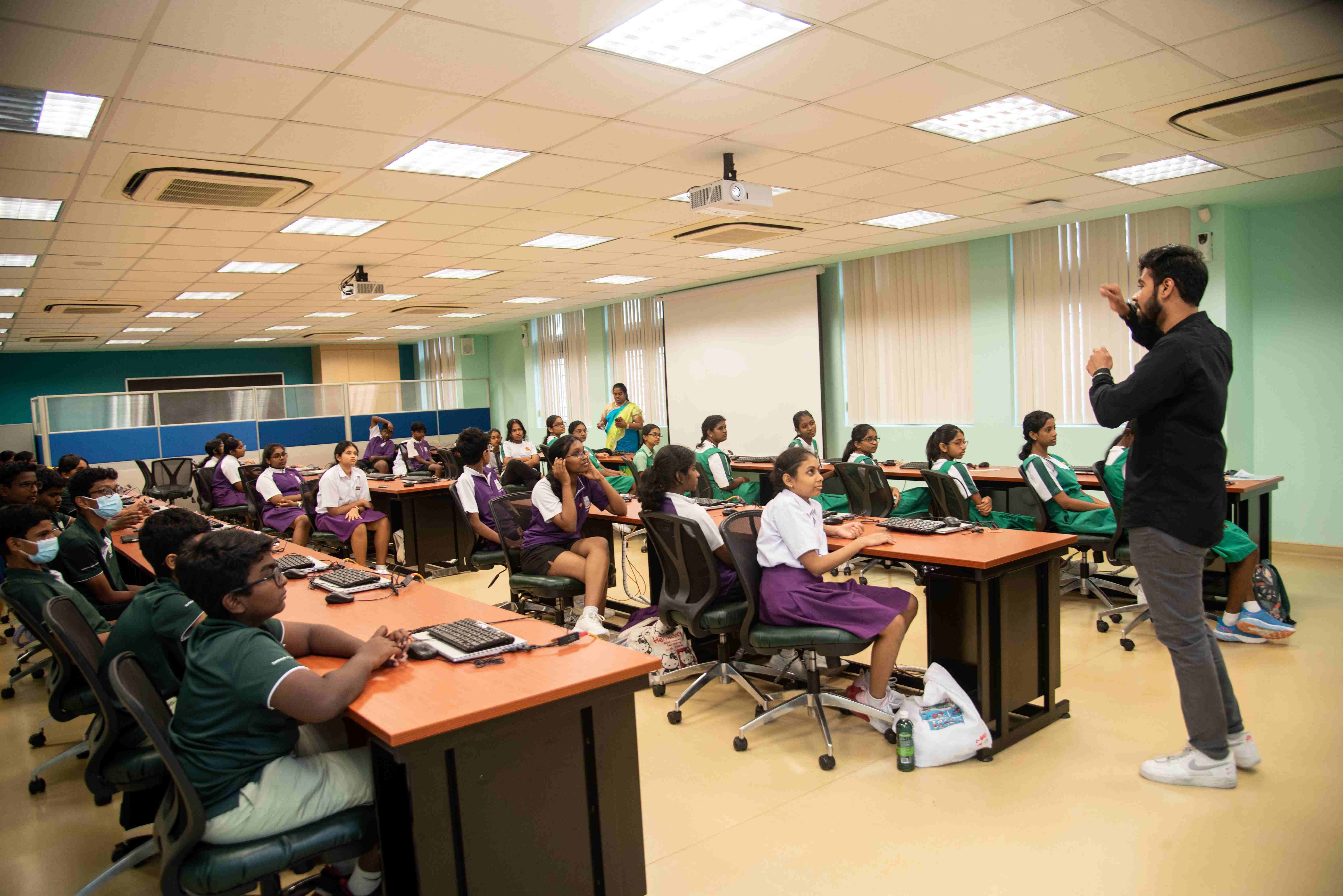 Instructor gestures while students in uniforms sit at computer desks in a classroom with ceiling projectors.