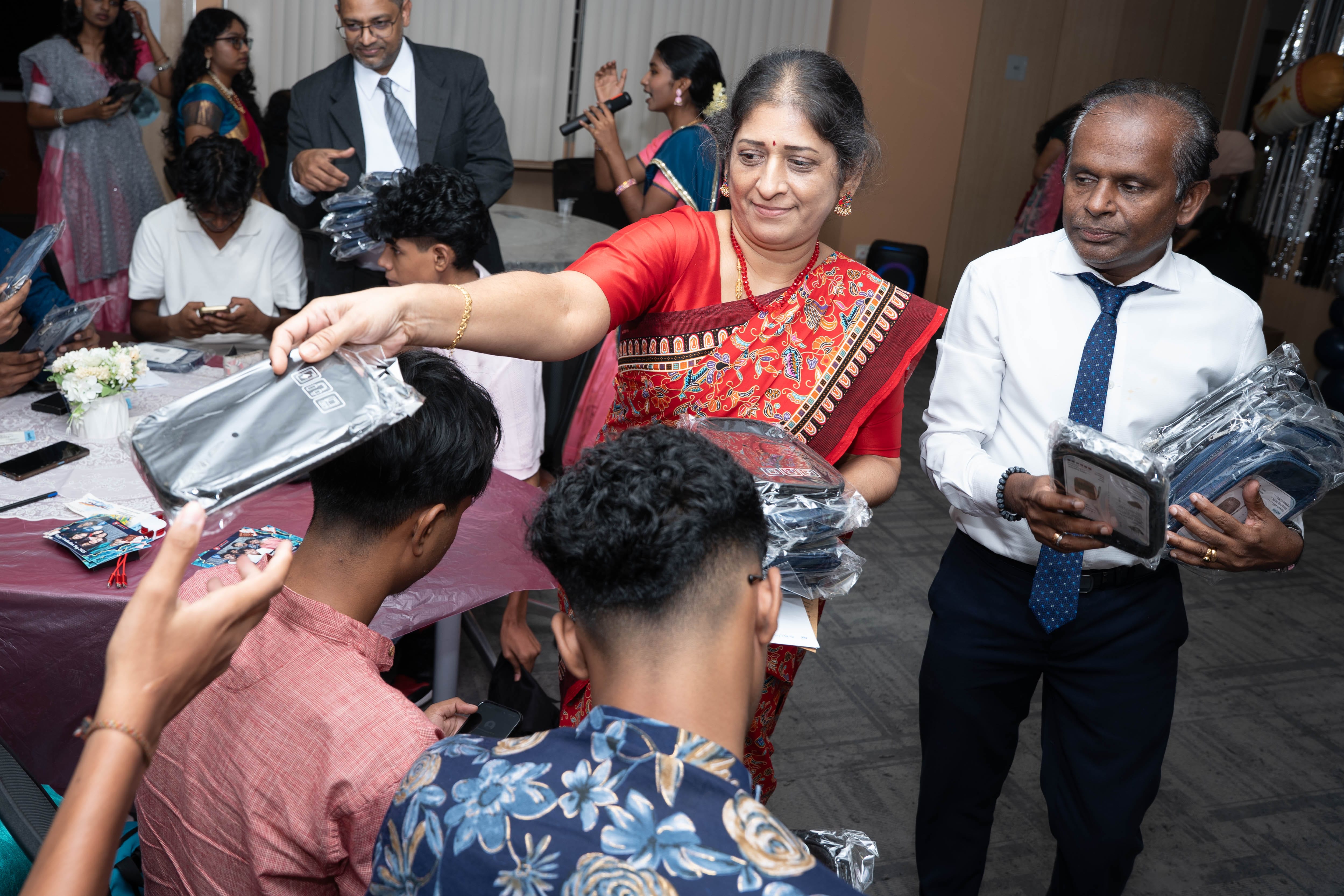 Woman in red sari hands packaged items to seated youths as man in tie holds more at a gathering.