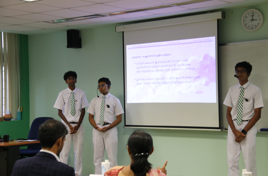 Three students in white uniforms and green ties present beside a projected slide in a classroom.