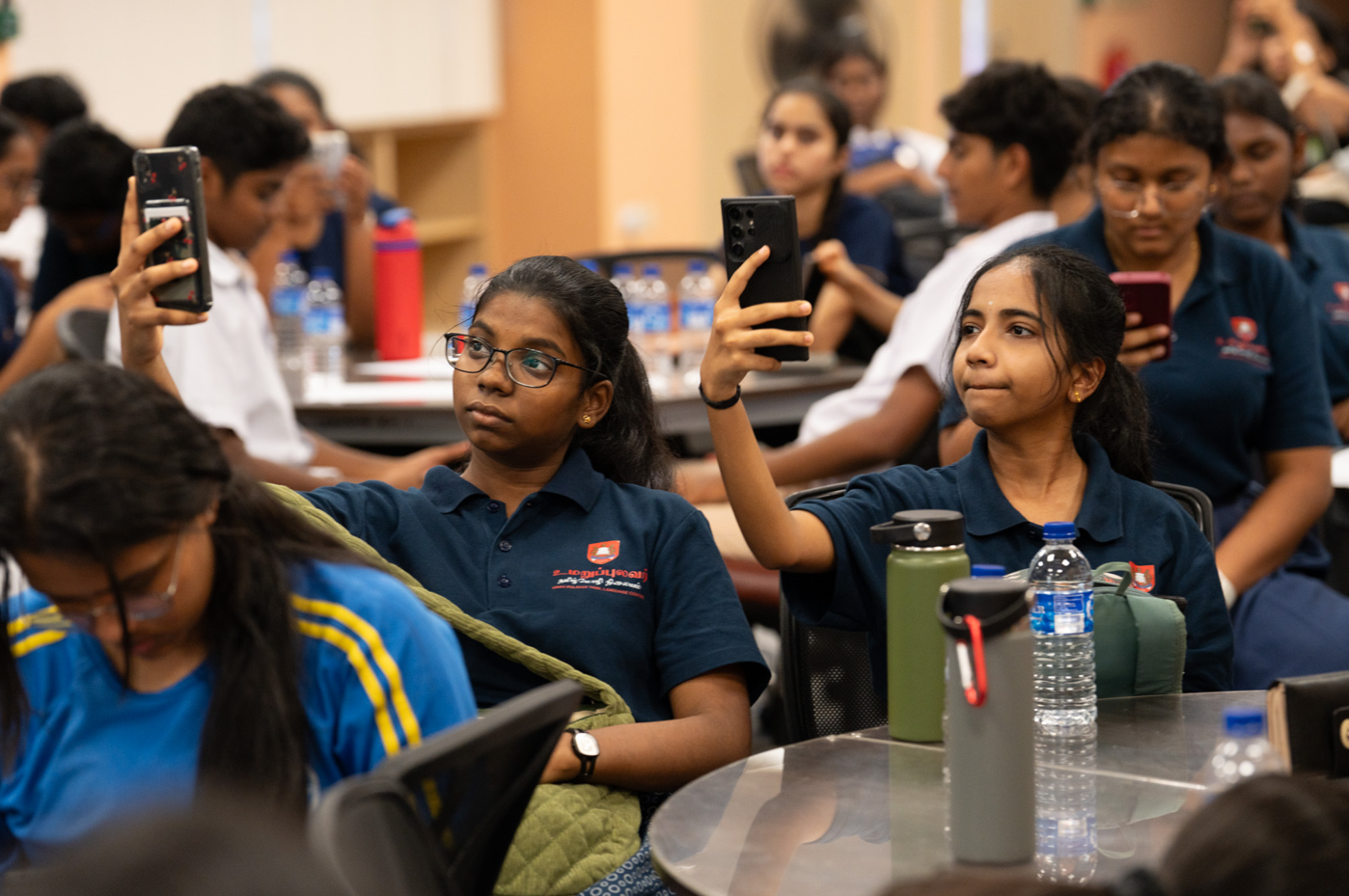 Two girls in navy polos sit at a table holding up phones, with other students and water bottles behind them.