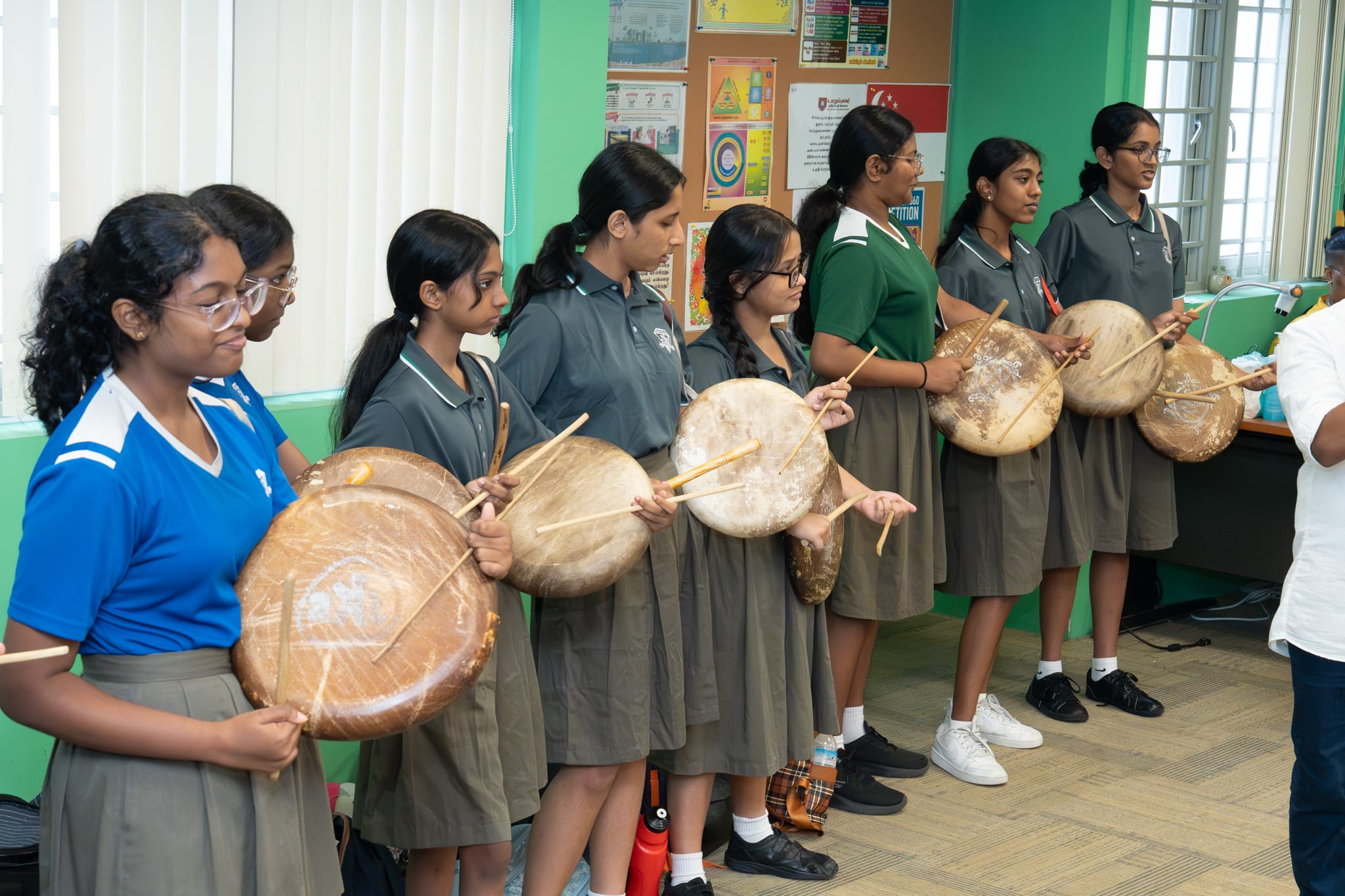 Schoolgirls stand in a line holding large frame drums and drumsticks in a classroom.