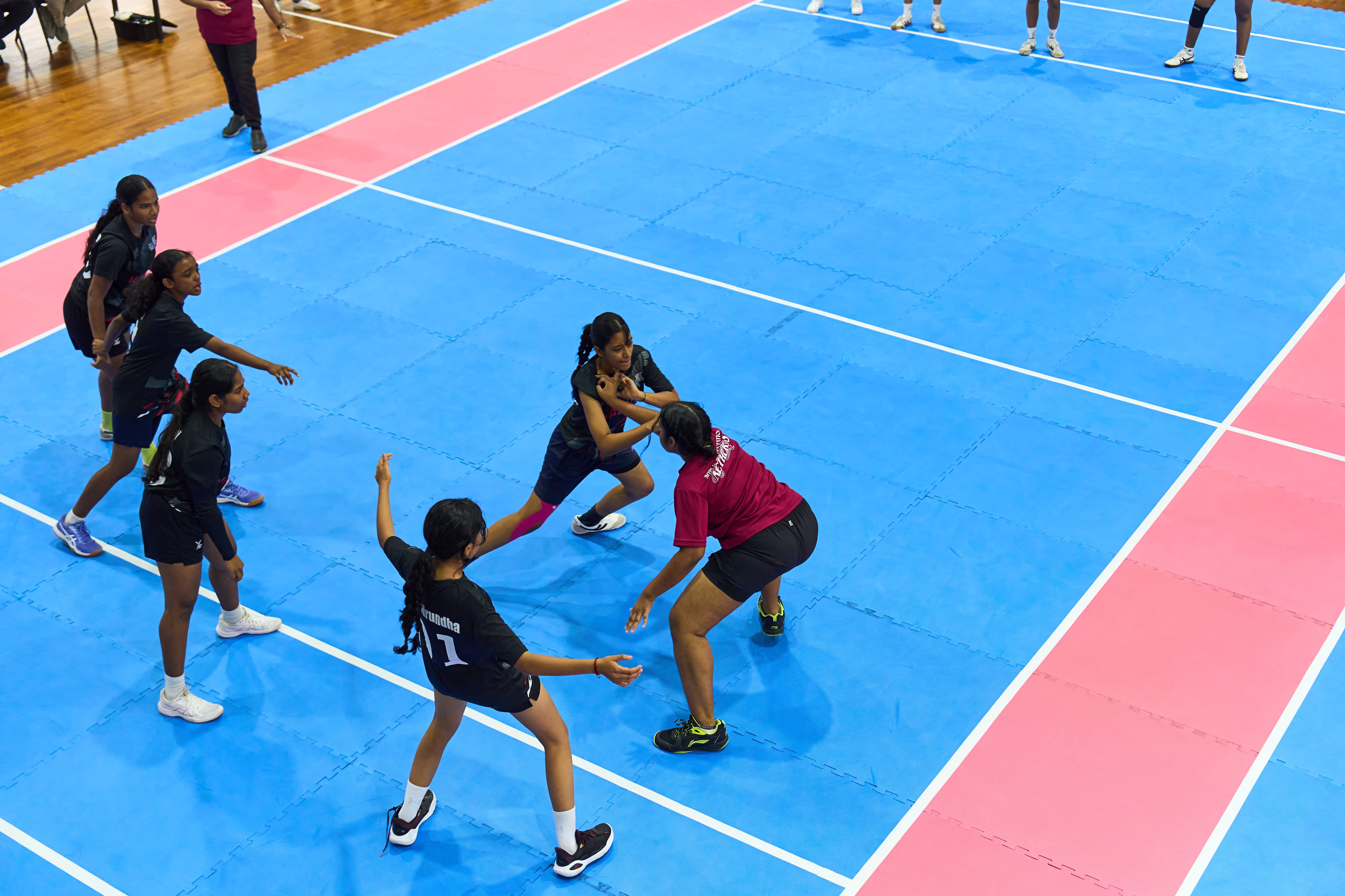 Women playing kabaddi on a blue mat court; one in red is grabbed by a defender as others surround.