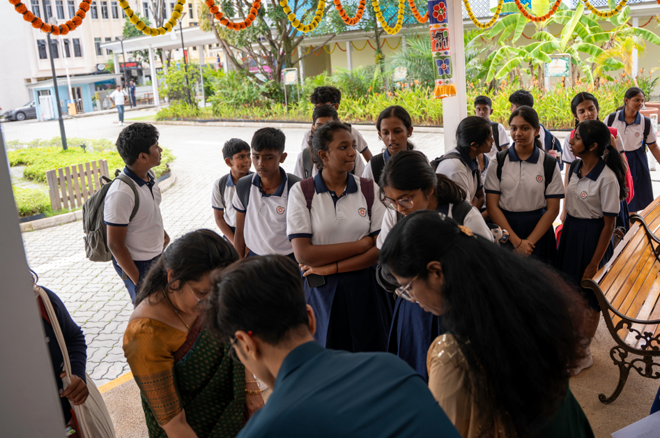Group of schoolgirls in white-and-navy uniforms gathered under colorful garlands near a campus entrance.