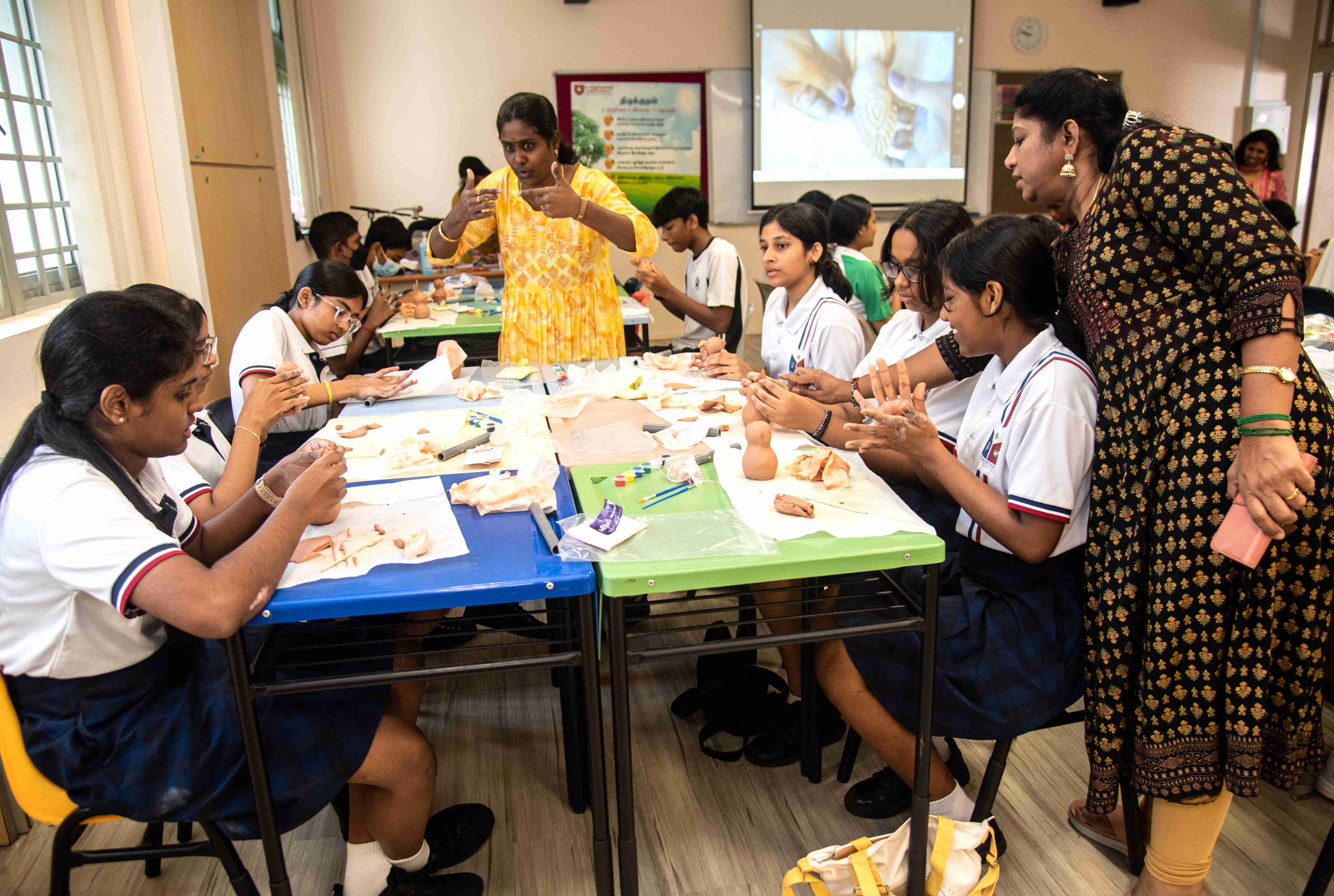 Students in uniforms sculpt clay at classroom tables while two teachers supervise.
