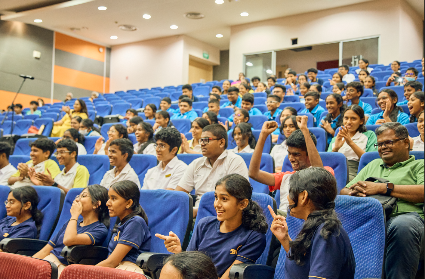 Students in an auditorium with blue seats clap and cheer, one raising both fists.