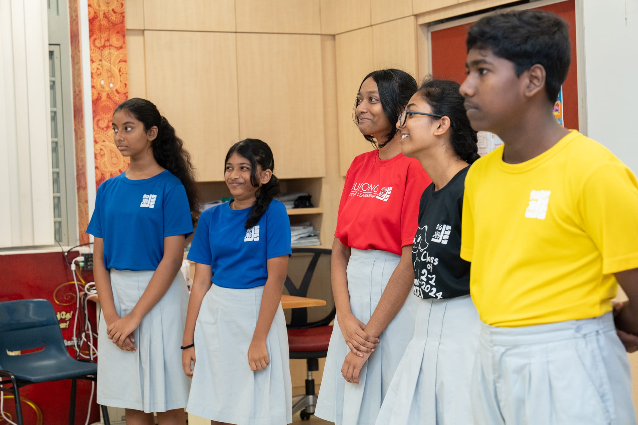 Five students in colorful T-shirts and light skirts stand in a classroom, looking to the right.