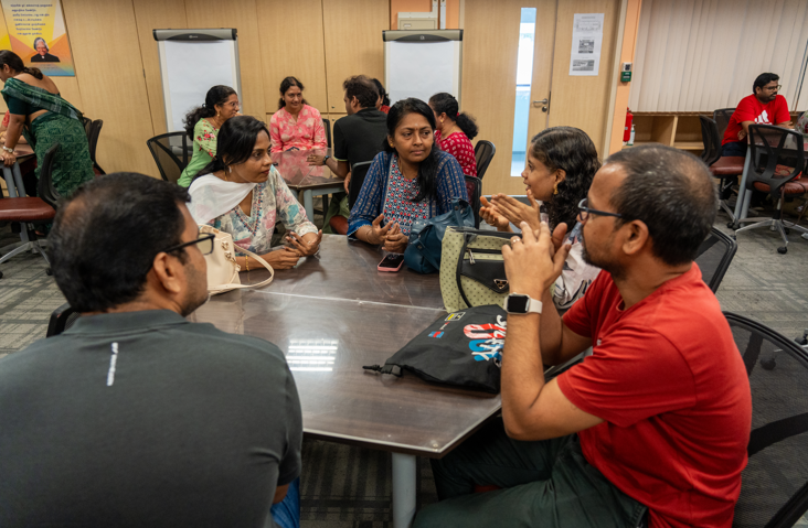 Group of people seated around a table in a meeting room, talking, with bags and a phone on the table.