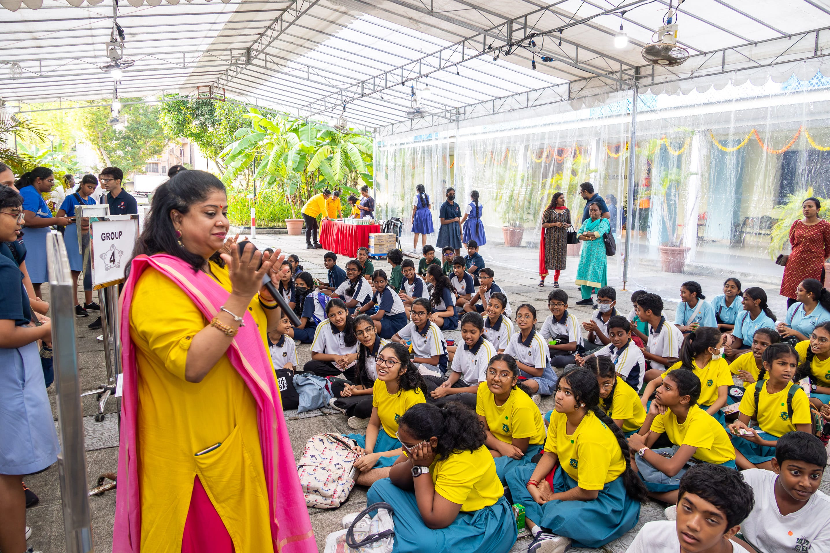 Woman in yellow with pink scarf speaks to seated schoolchildren in uniforms under a covered outdoor area.
