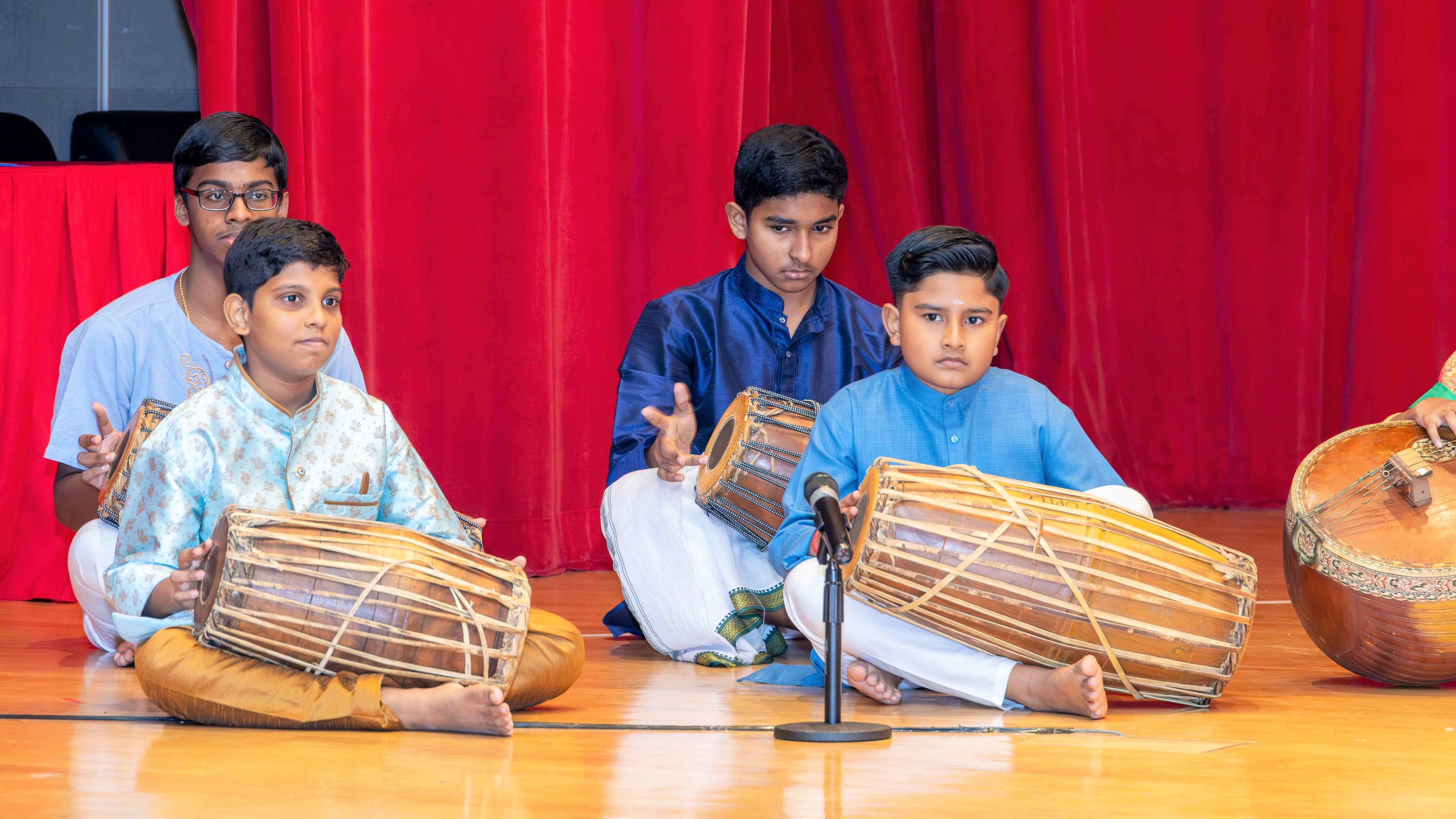 Four boys seated on stage play barrel drums in front of a red curtain, with a microphone in front.