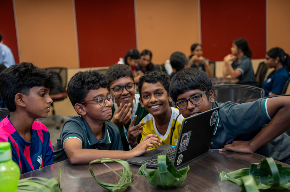 Five kids gathered around a laptop at a table, smiling and making a peace sign in a classroom.