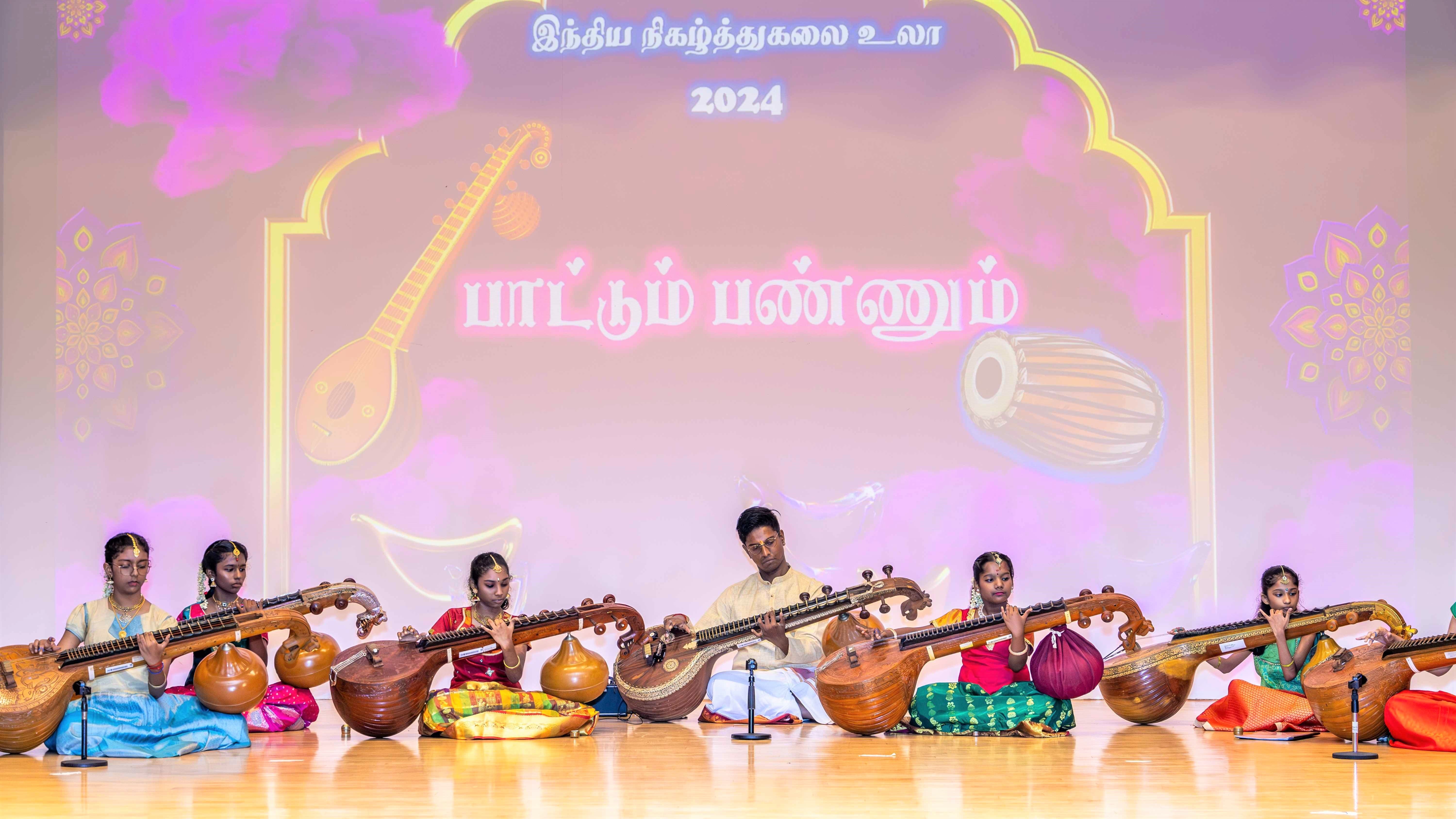 Six seated musicians on stage play veenas; Tamil text and 2024 on backdrop with veena and drum icons.