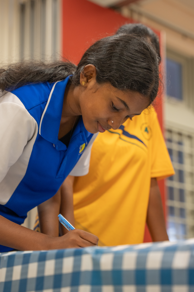 Woman in blue shirt leans over a table, writing with a pen; person in yellow shirt stands behind.