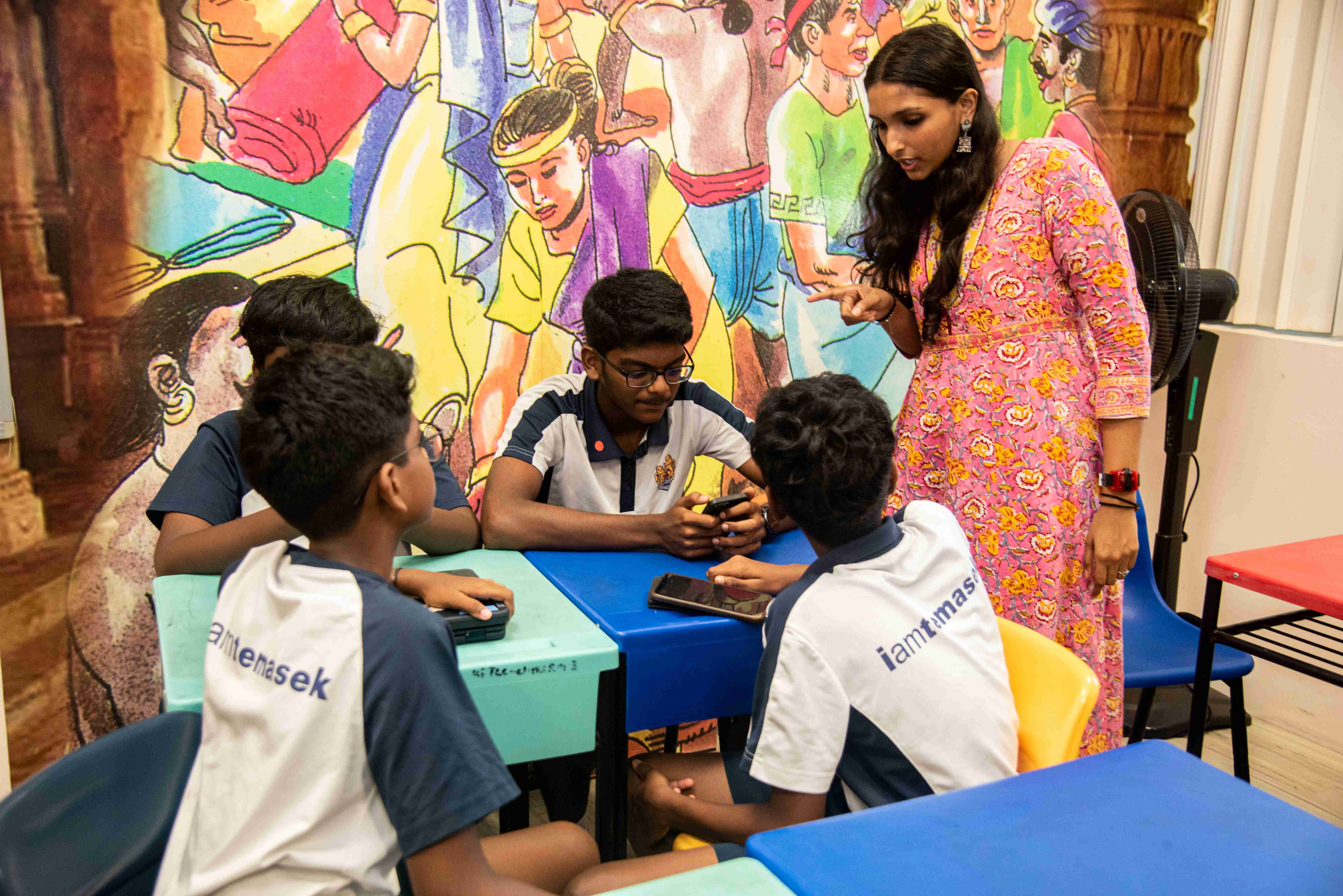 Teacher in pink dress points while boys in uniforms sit at tables using phones, mural behind them.