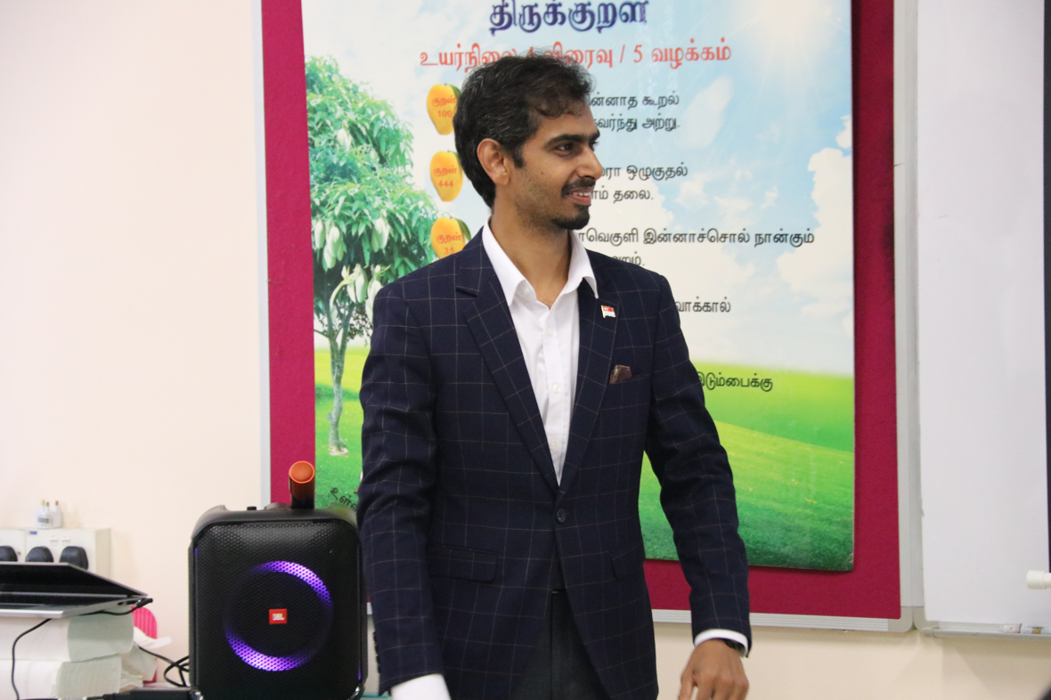 Man in a checkered suit stands indoors beside a lit speaker, with a Tamil poster and tree behind him.