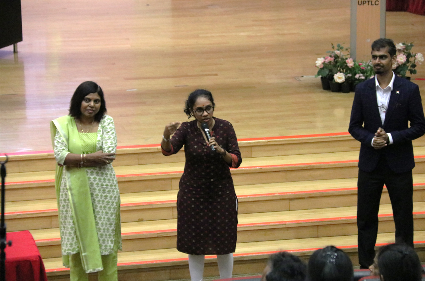 Woman speaks into a microphone on stage steps, flanked by a woman in green and a man in a suit.