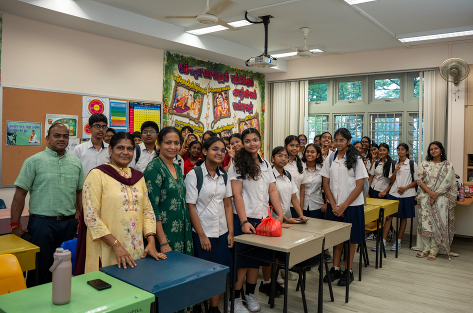 Group of students and teachers posing in a classroom with desks, wall posters, and large windows.