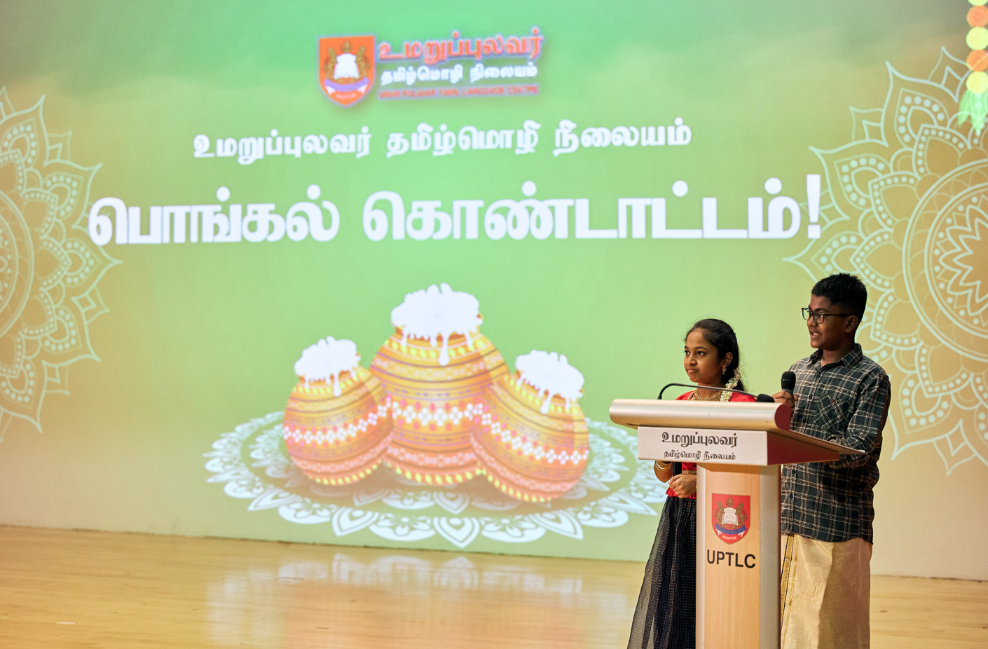 Two students speak at a podium with microphone on stage before a green Tamil Pongal festival backdrop.