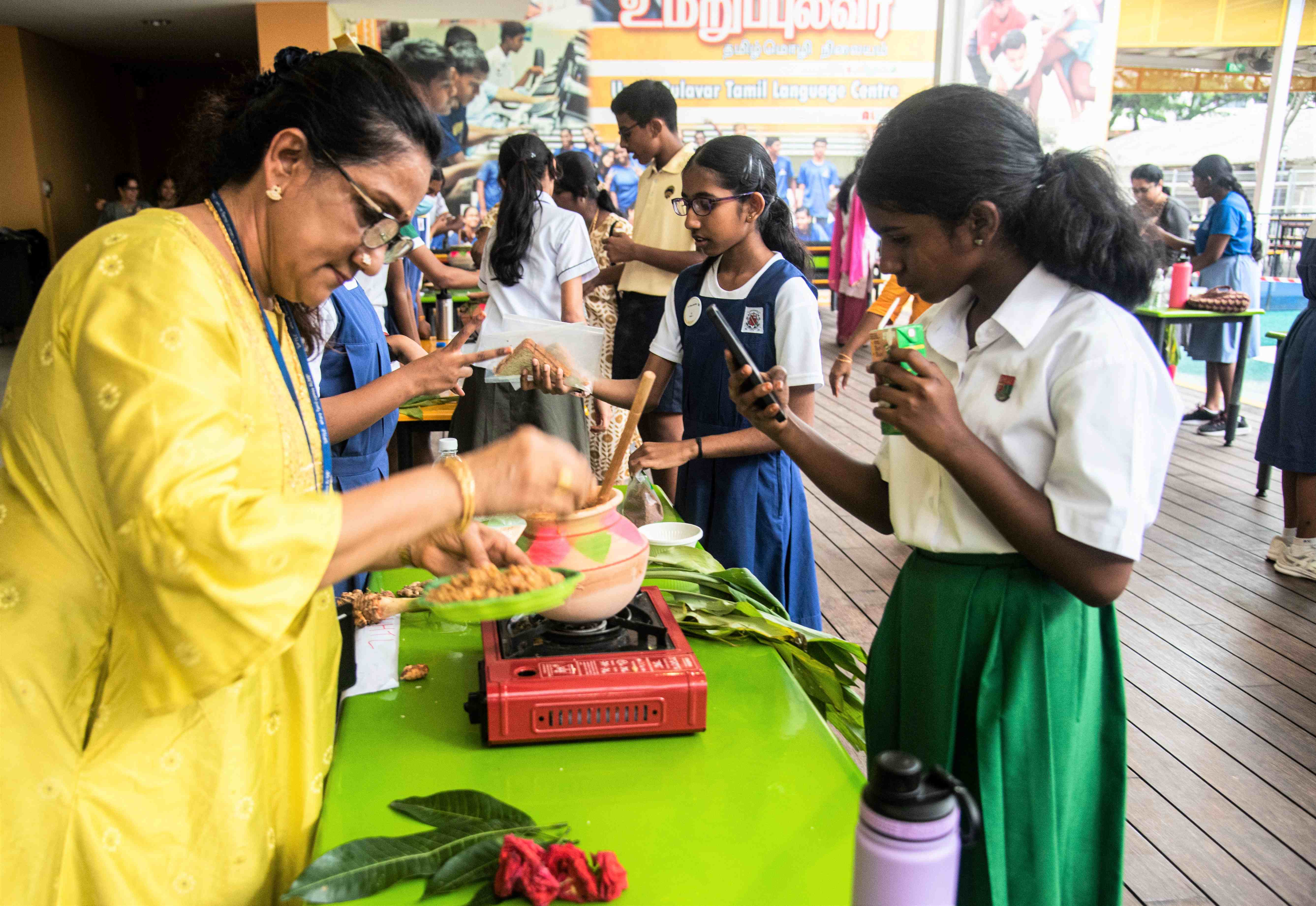 Woman in yellow serves food at a table with a pot on a burner as schoolgirls watch and taste samples.