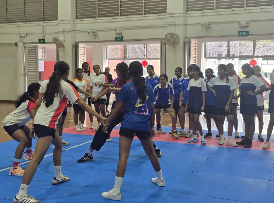 Girls in sports uniforms watch a coach demonstrate a drill on blue mats in an indoor gym.